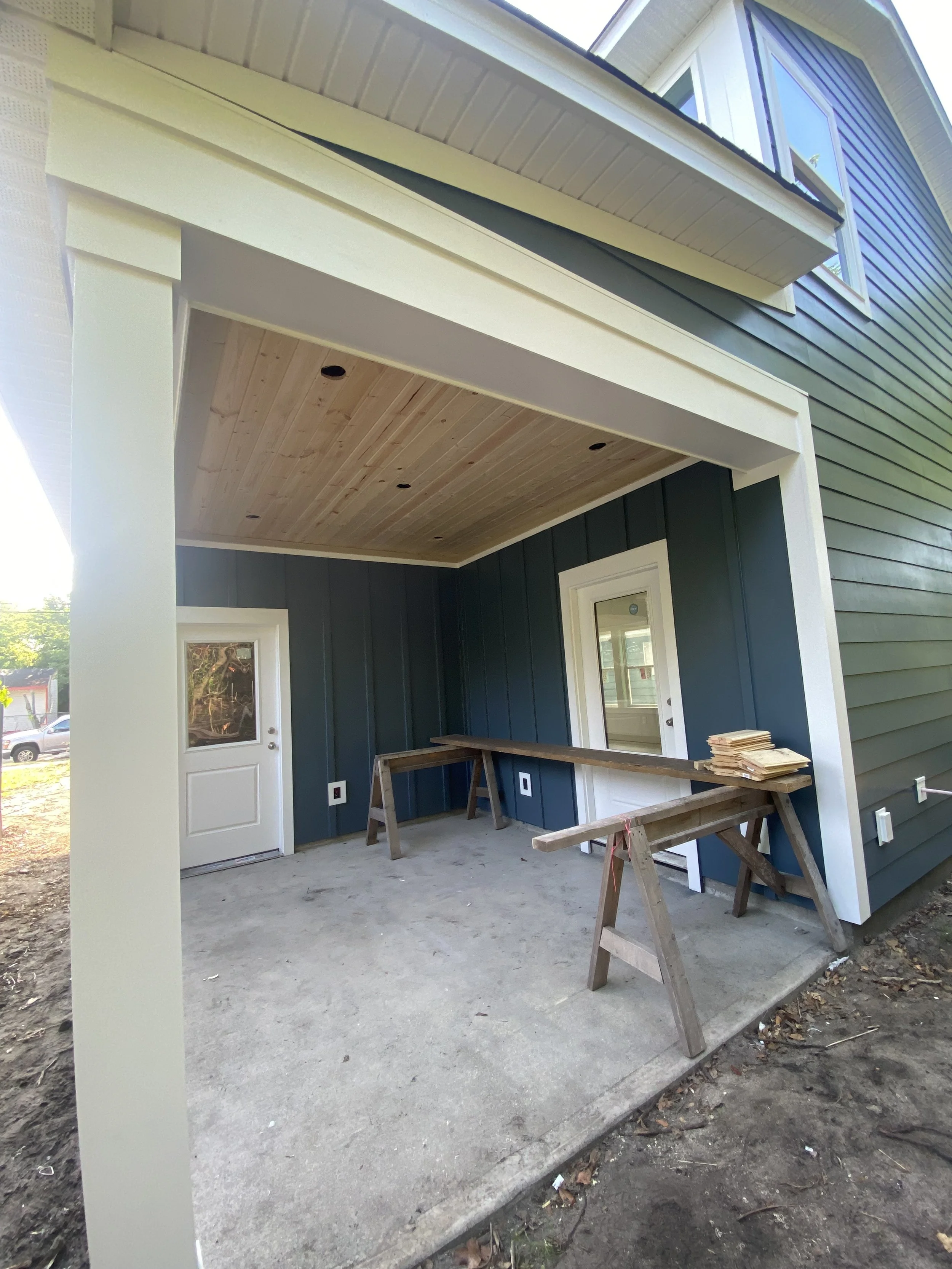 Under-construction patio with dark green siding, a blue interior wall, a door, a window, a wood-paneled ceiling with recessed lights, and construction tools and materials, including a sawhorse and stacked wood boards.