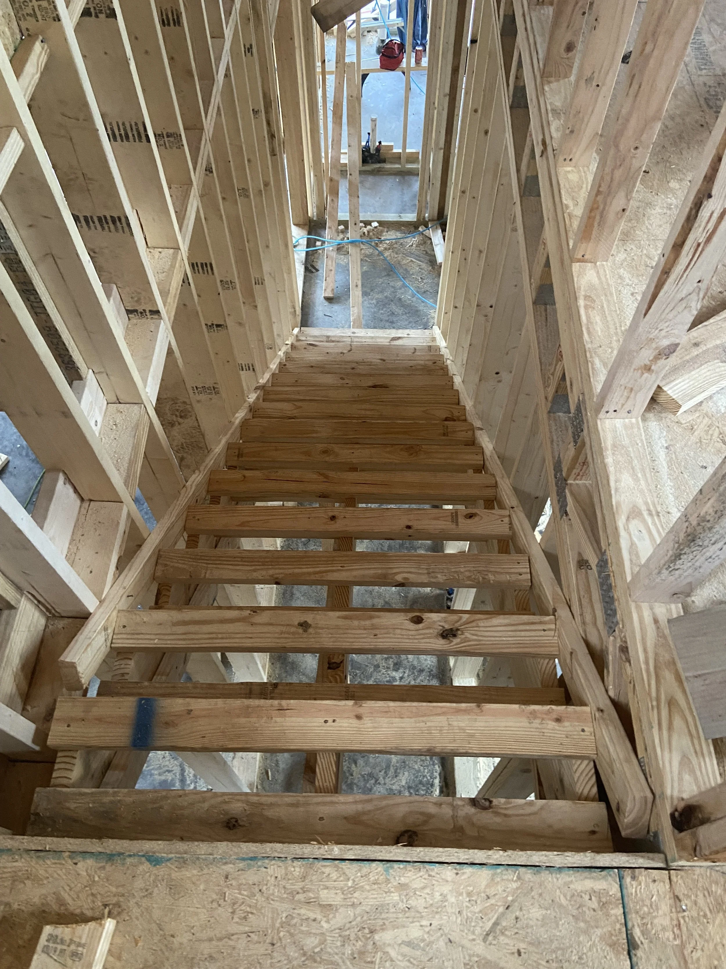 Wooden staircase under construction inside a framed building, showing the steps and surrounding wall studs.