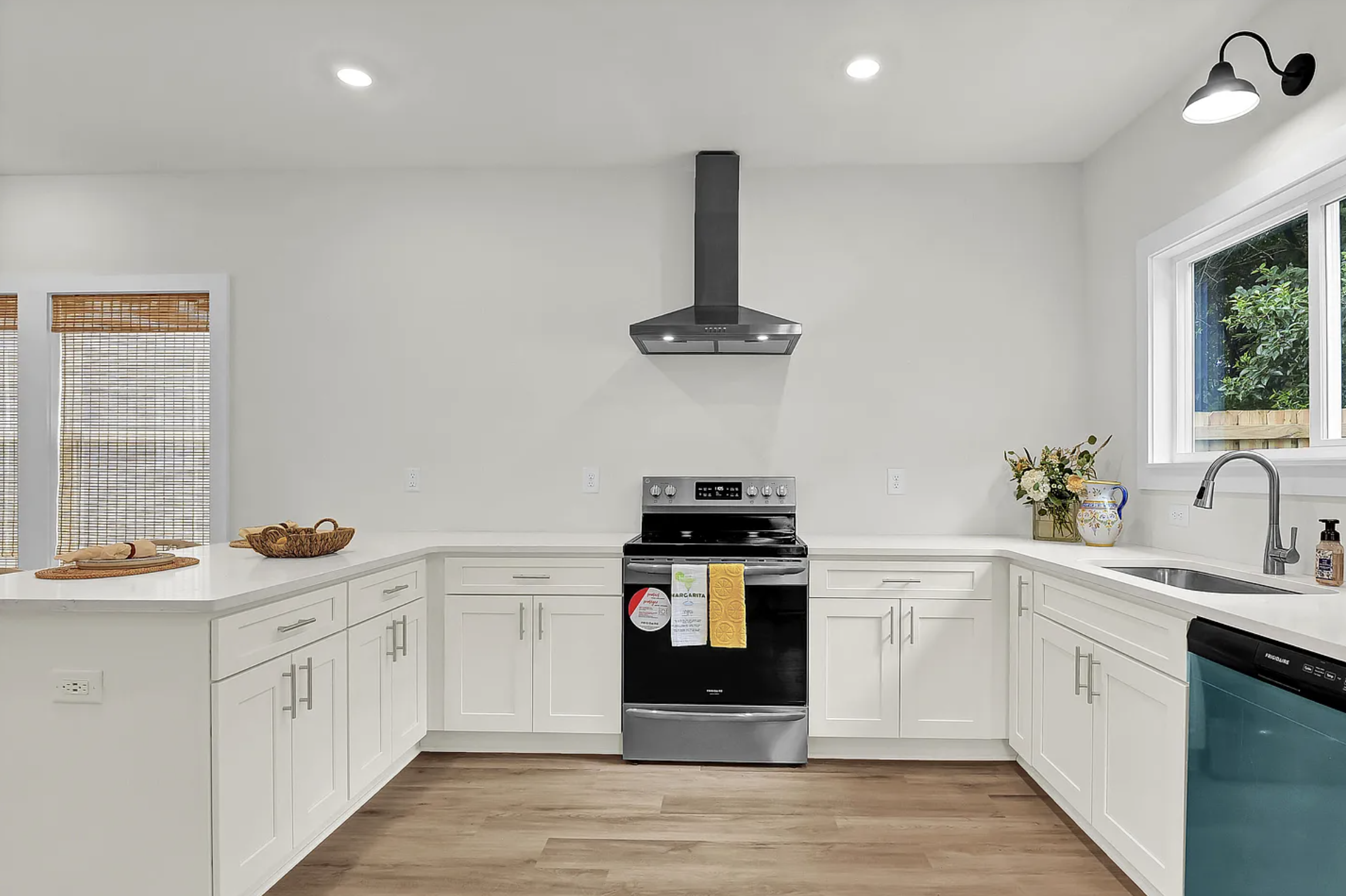 Modern kitchen with white cabinets, stainless steel stove and range hood, black dishwasher, window with view of trees, and wooden floor.