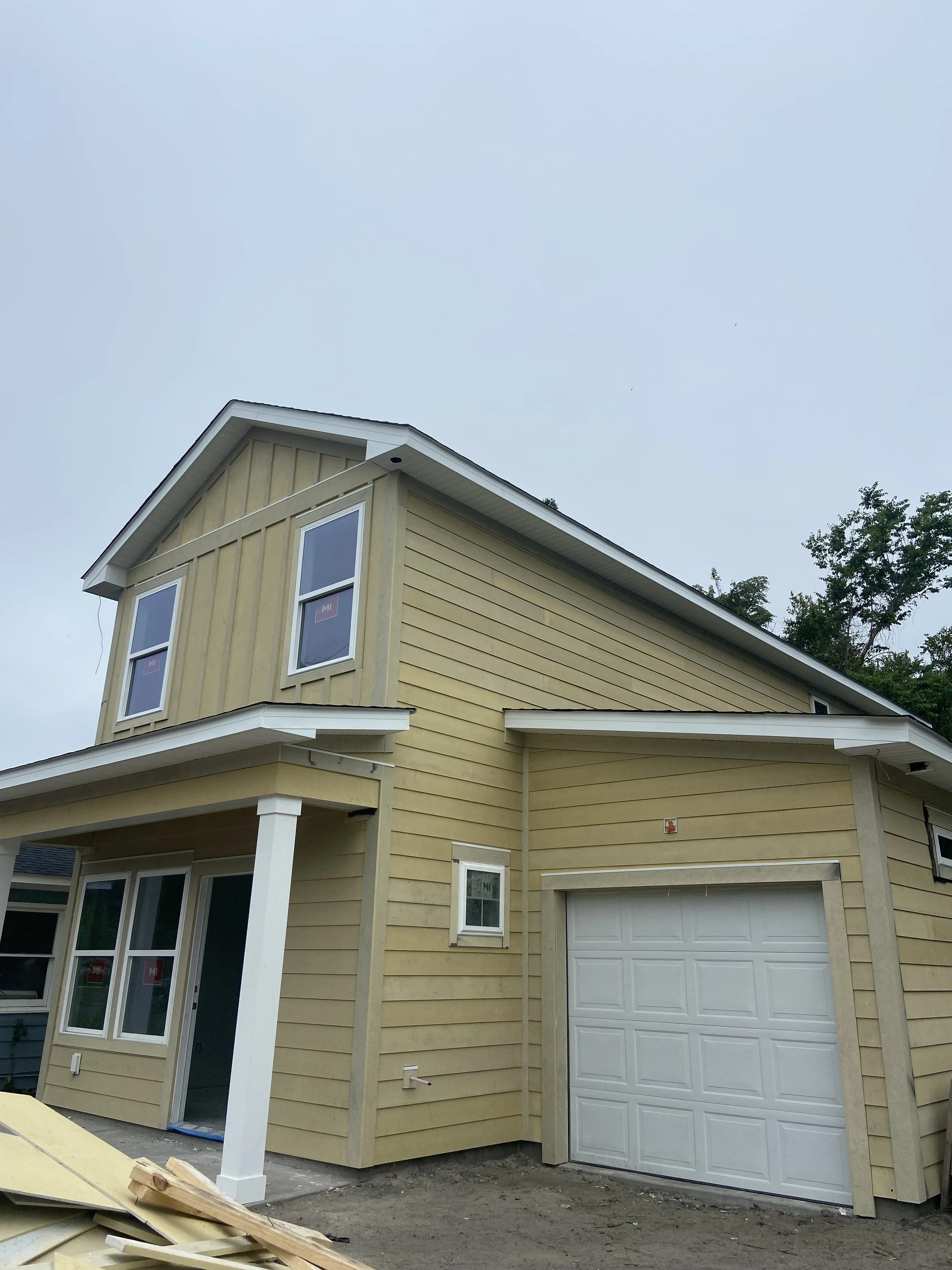 A yellow two-story house under construction with white trim, black roof shingles, a garage door, and windows with purple stickers, surrounded by dirt and building materials.