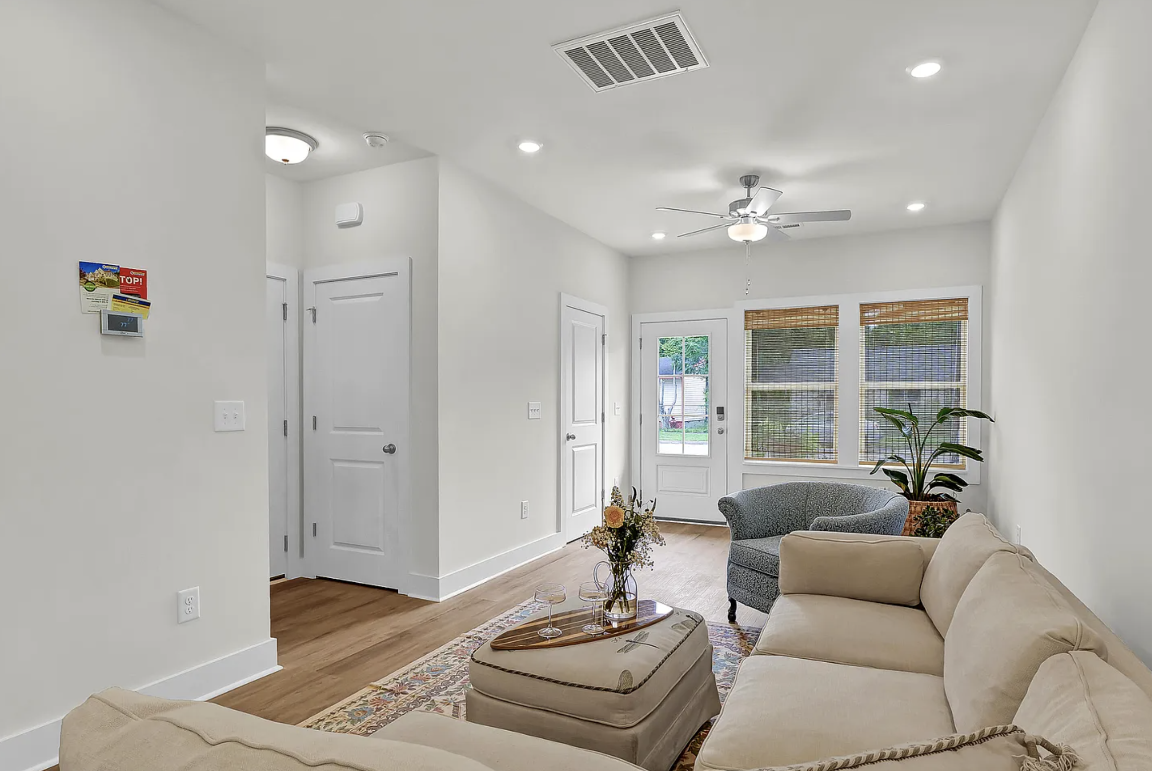 Living room with white walls, hardwood floors, beige sofa, blue armchair, large windows with bamboo blinds, potted plant, ceiling fan, and pendant lighting.