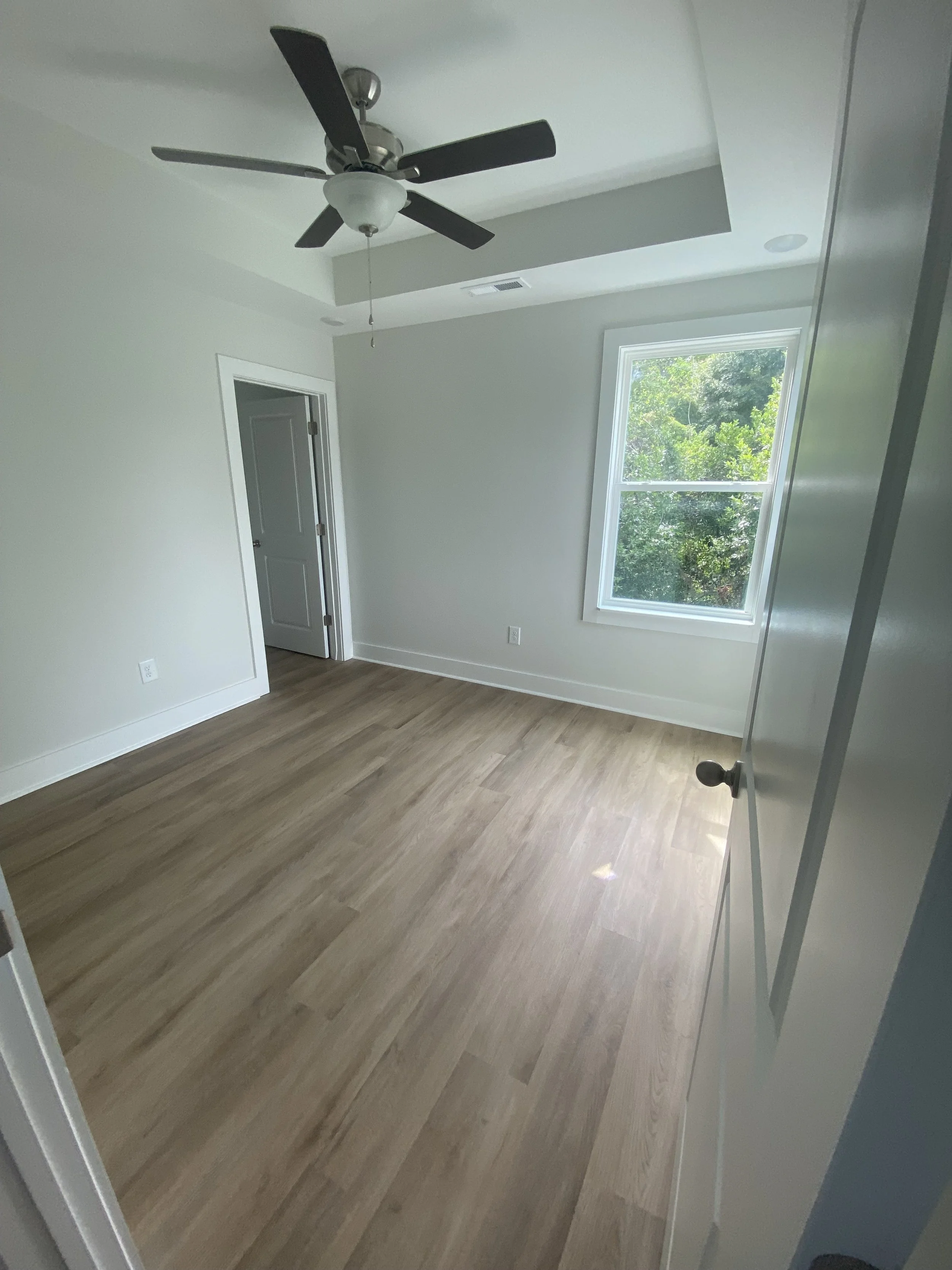Empty room with hardwood flooring, a ceiling fan, a large window with greenery outside, and a partly open door.