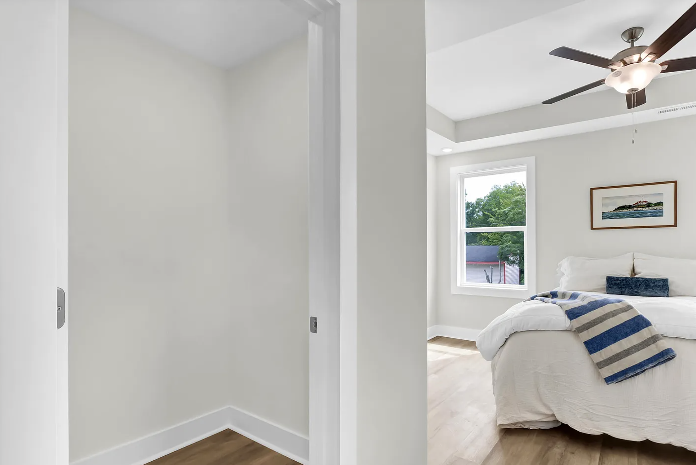 View of a bedroom with a bed, window, ceiling fan, and artwork on the wall; adjacent space with white walls and wood floor.
