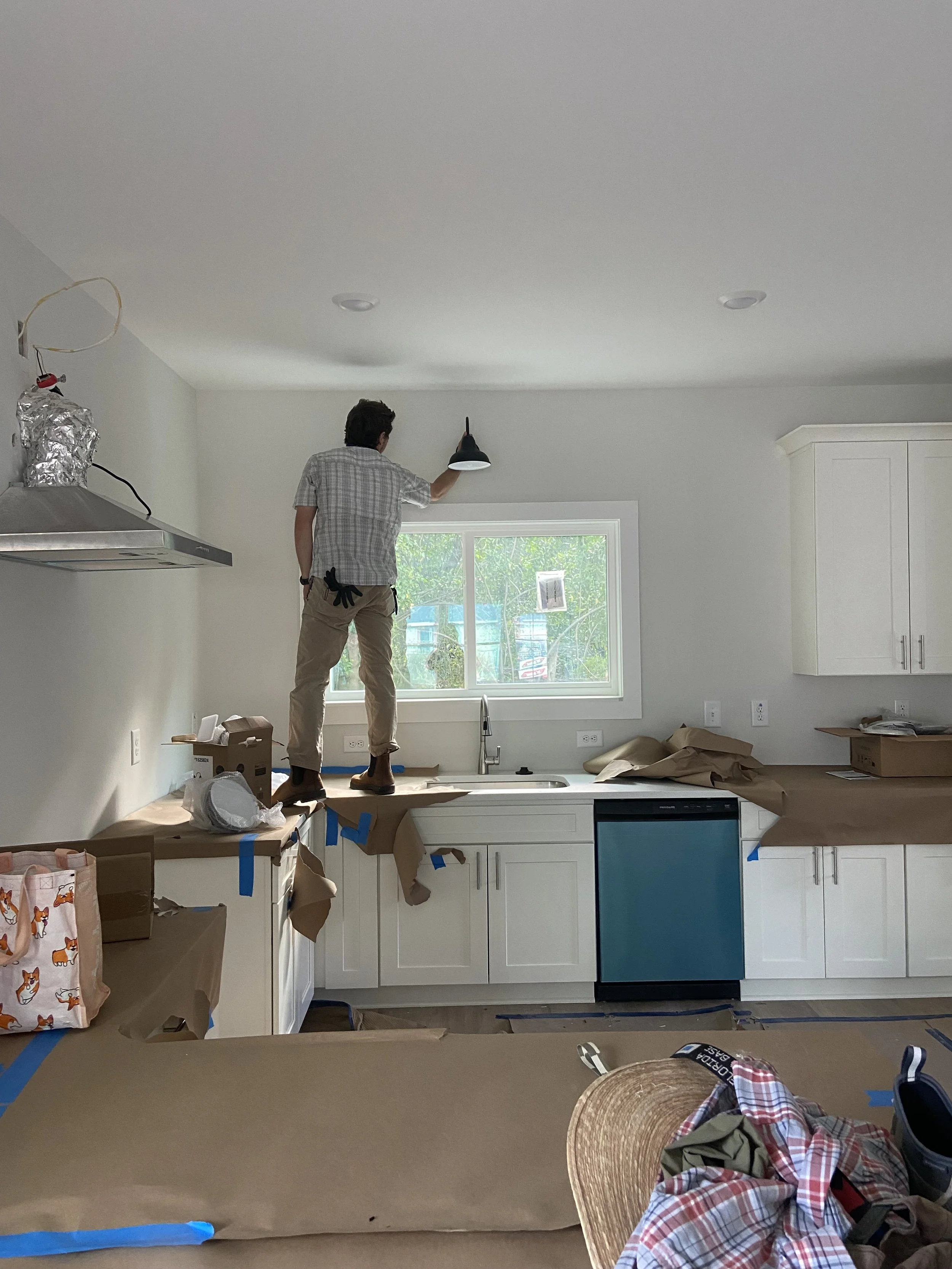 Person standing on kitchen counter installing a ceiling light fixture, with kitchen cabinets, window, and dishwasher visible in the background, while wearing casual clothing.