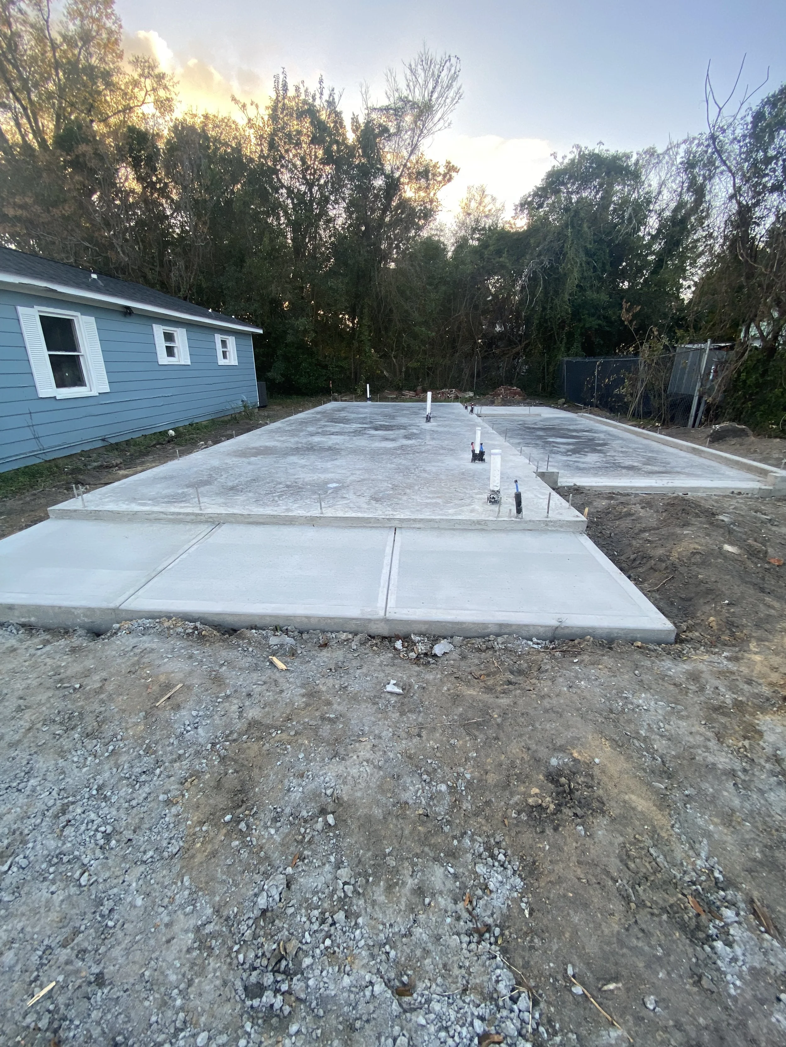 Photo of a residential construction site showing freshly poured concrete slab foundation with pipes and fixtures protruding, surrounded by dirt and soil, with a blue house and trees in the background, and a partly cloudy sky overhead.