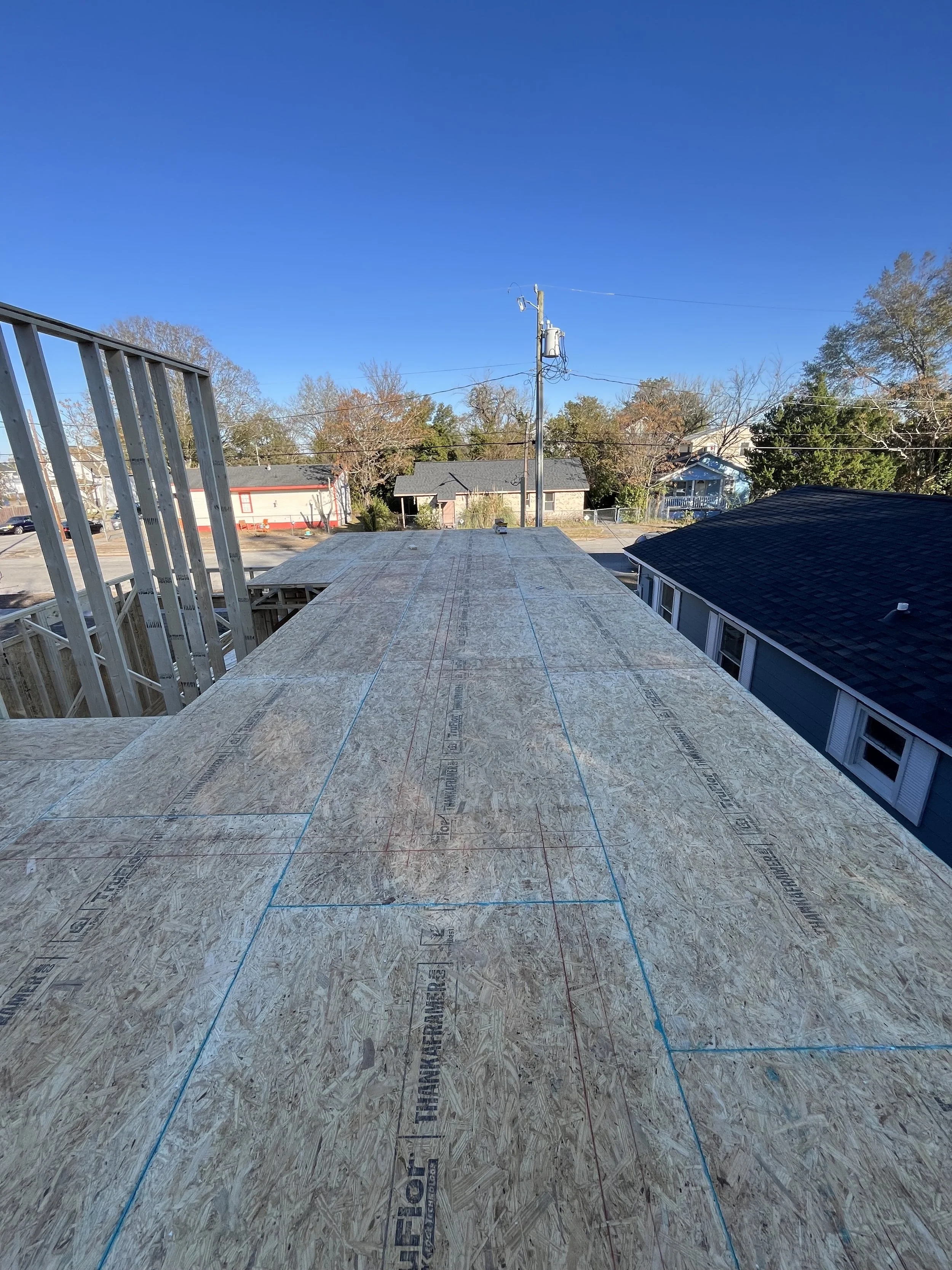 View of a construction site with wooden plywood flooring and framing on a building, overlooking a neighborhood with houses, trees, and a utility pole under a clear blue sky.