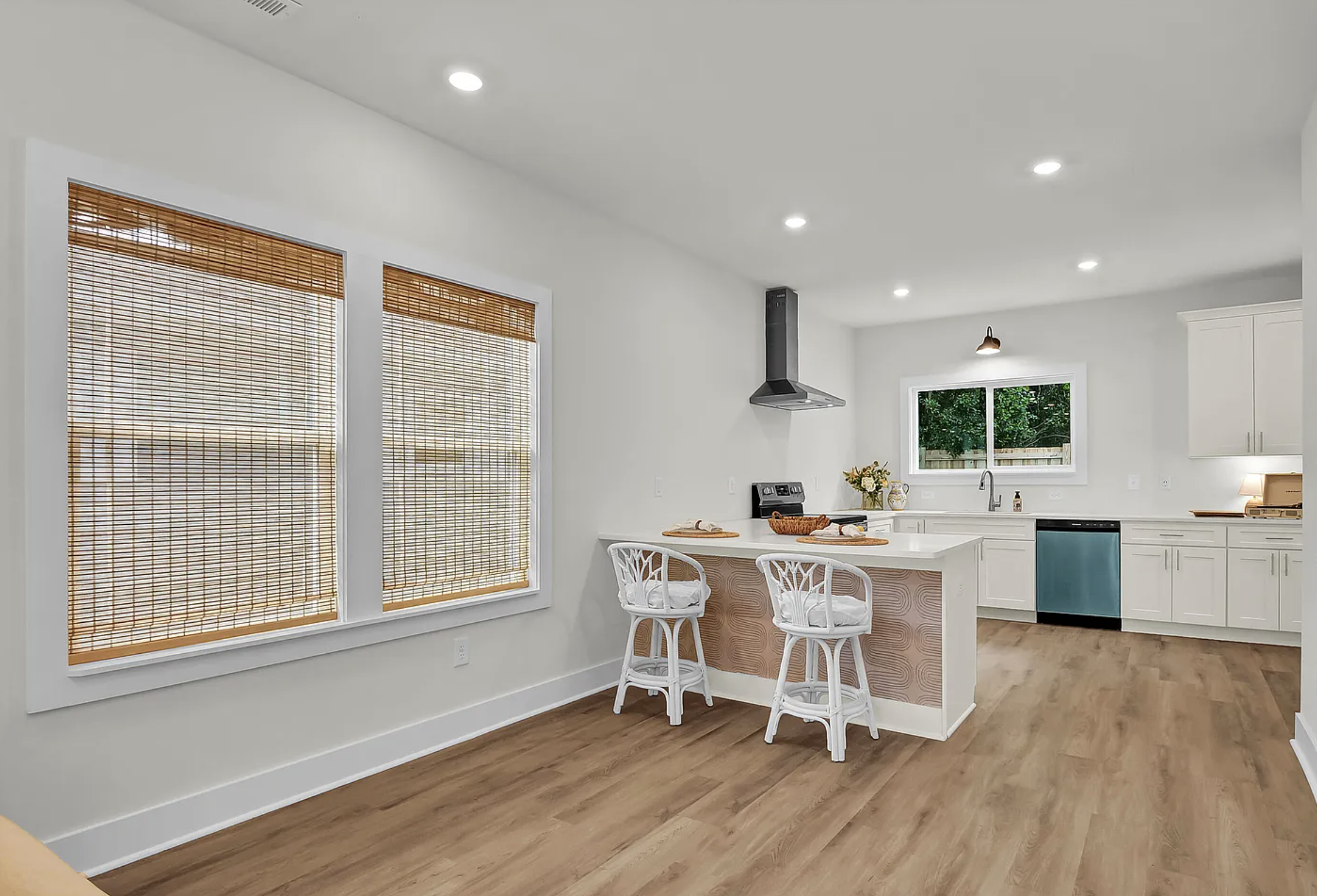 Bright, modern kitchen with white cabinets, wooden flooring, large windows with bamboo blinds, and a kitchen island with two white chairs.