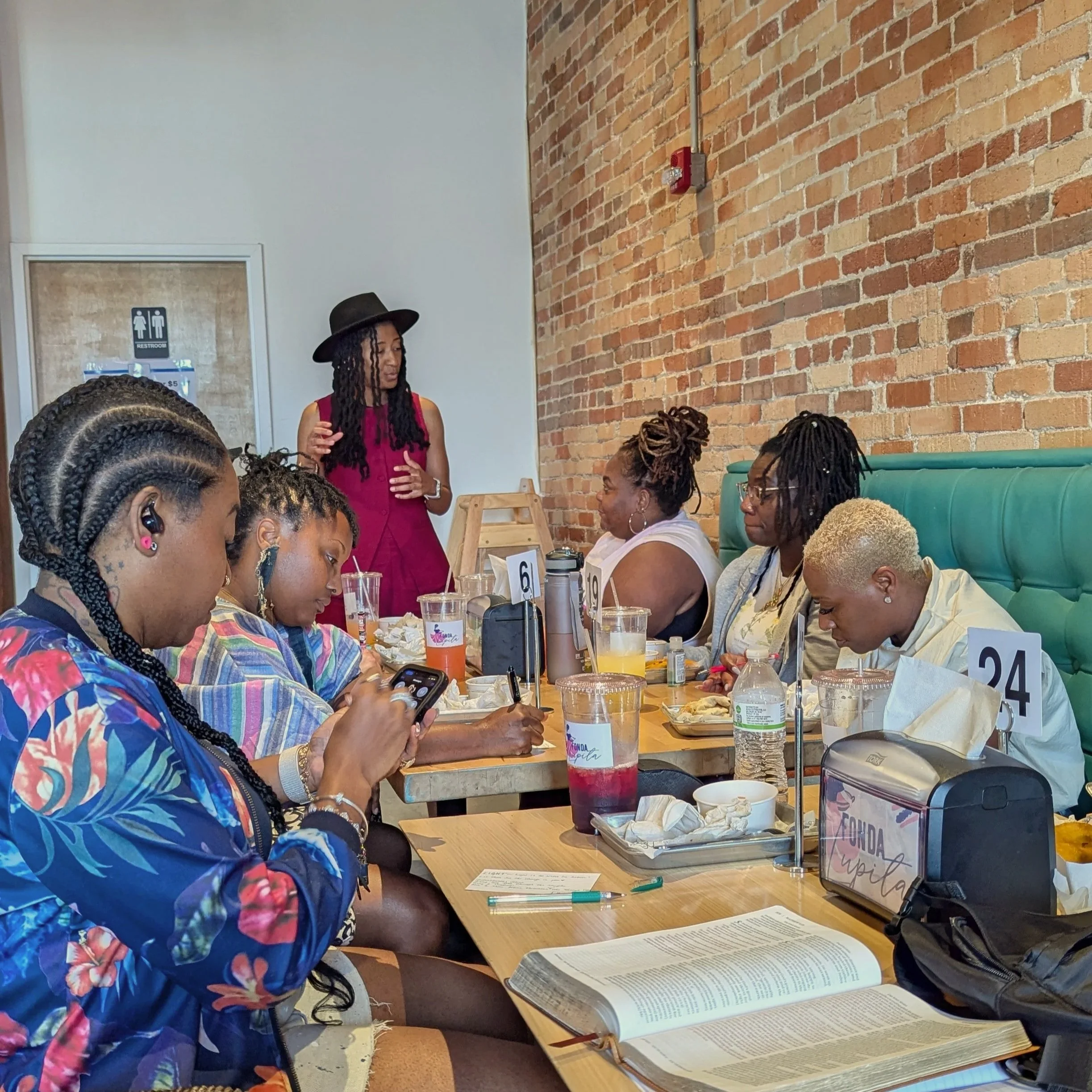 Group of women sitting at a restaurant table, some looking at their phones, with one woman standing and speaking, against a brick wall background.