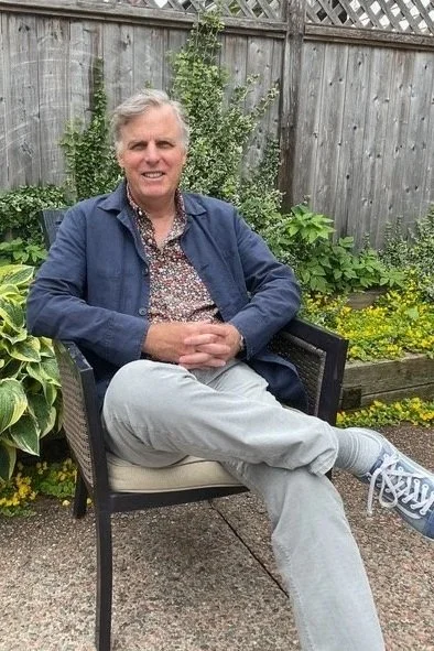 A man with gray hair sitting outdoors in a garden, smiling, wearing a patterned shirt, a dark jacket, light pants, and sneakers, with a wooden fence and green plants in the background.
