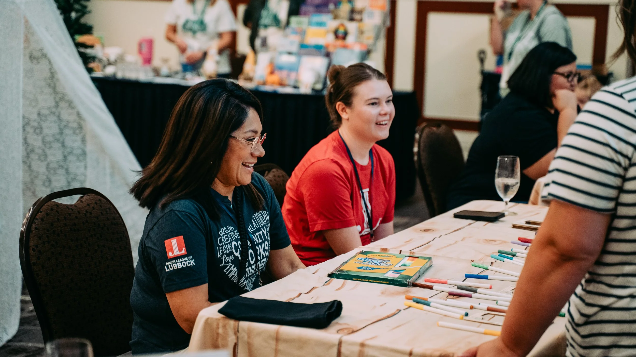 Two women sitting at a table with colorful markers, smiling and talking to a person standing in front of them at the Lubbock Book Festival.