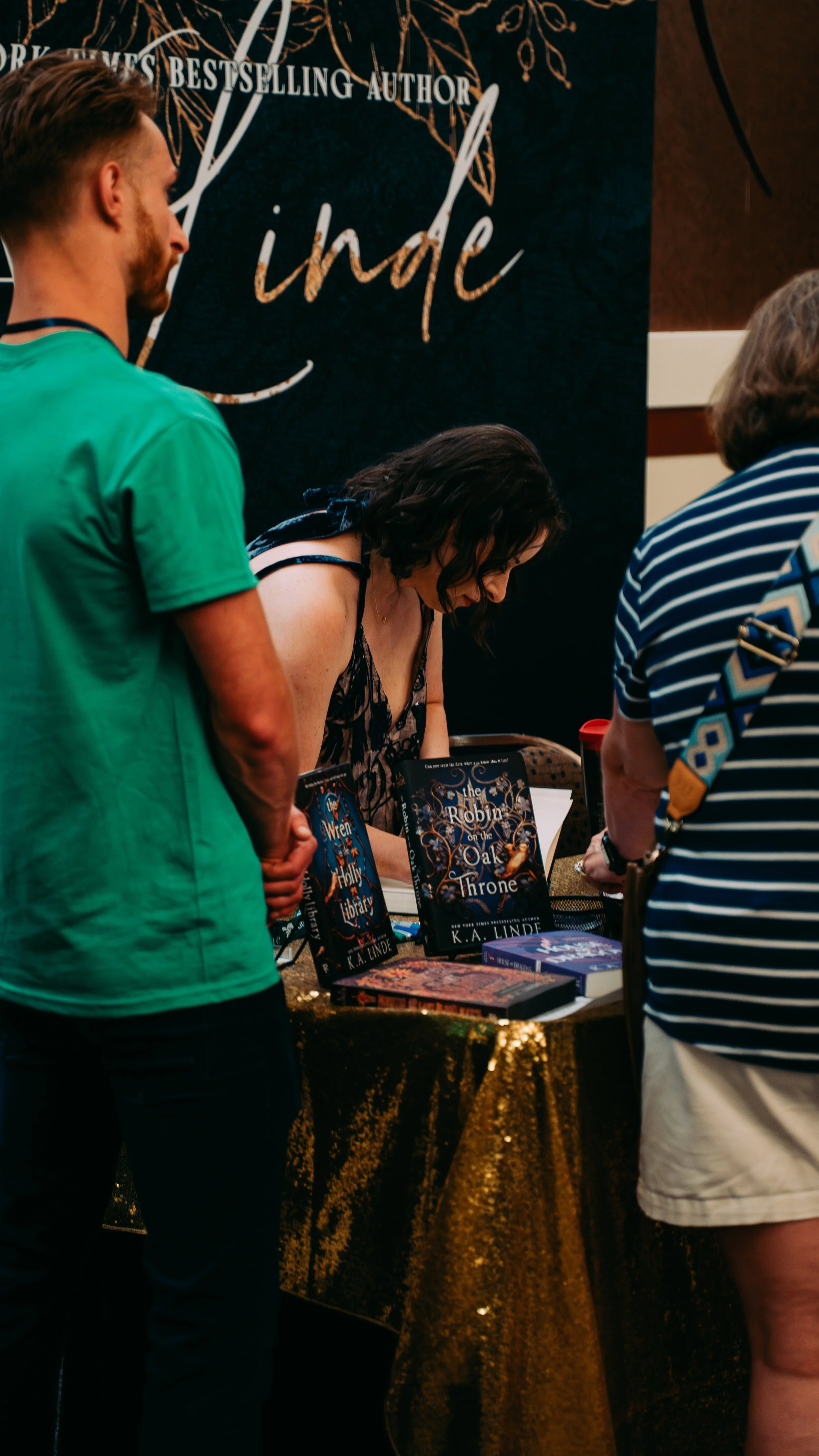 Author K. A. Linde signing books at a book signing event, with books displayed on a table covered with a gold sequin tablecloth.