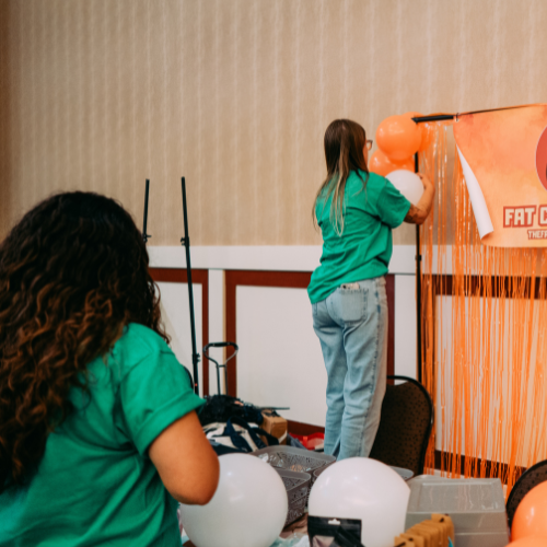 A woman in a green shirt decorating an author's background banner with orange balloons, while another woman in a green shirt with curly hair is preparing the balloons.