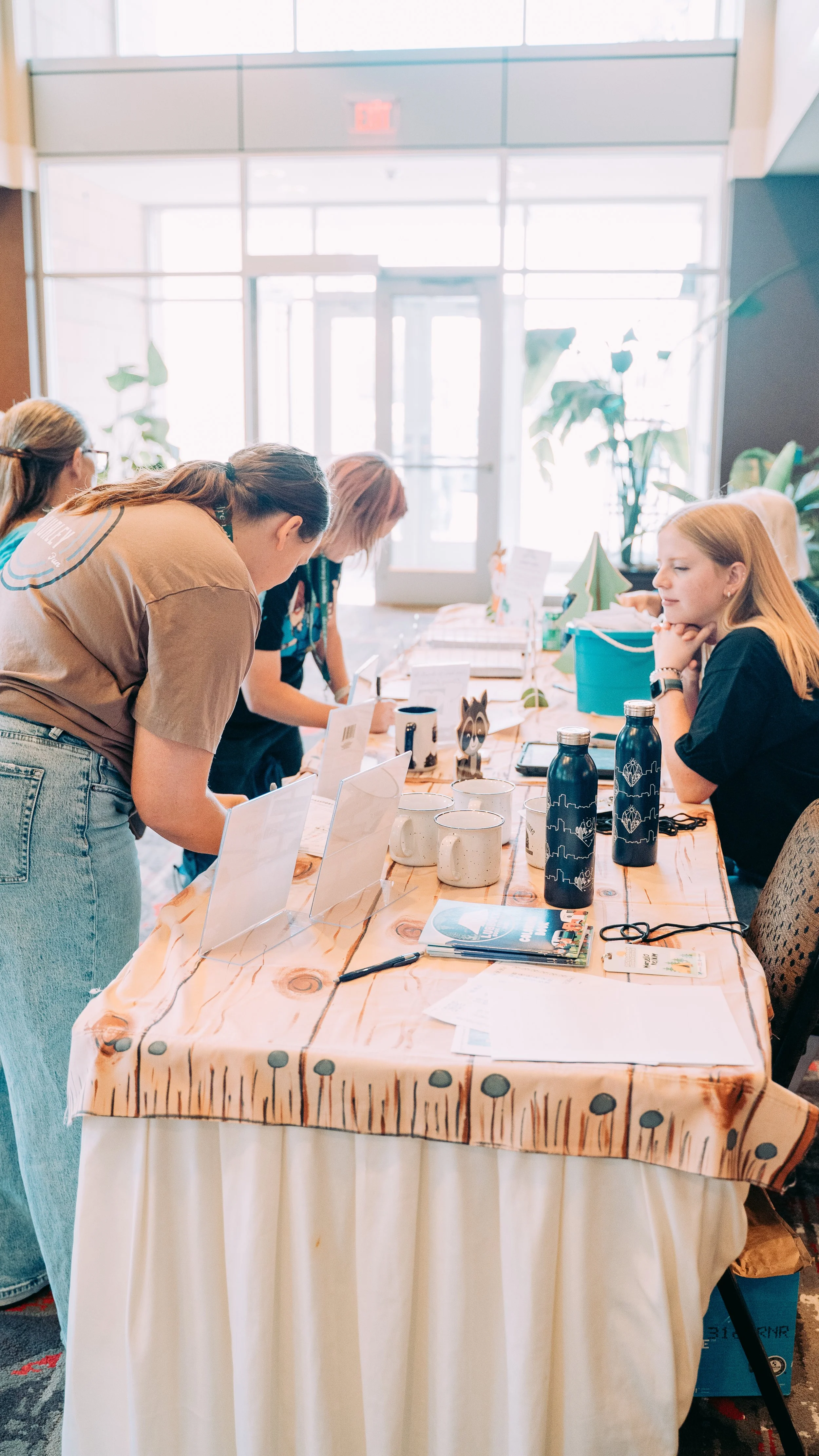 People browsing items at a registration or info table in a well-lit indoor space with large windows, potted plants, and various items like mugs, water bottles, brochures, and papers on the table.