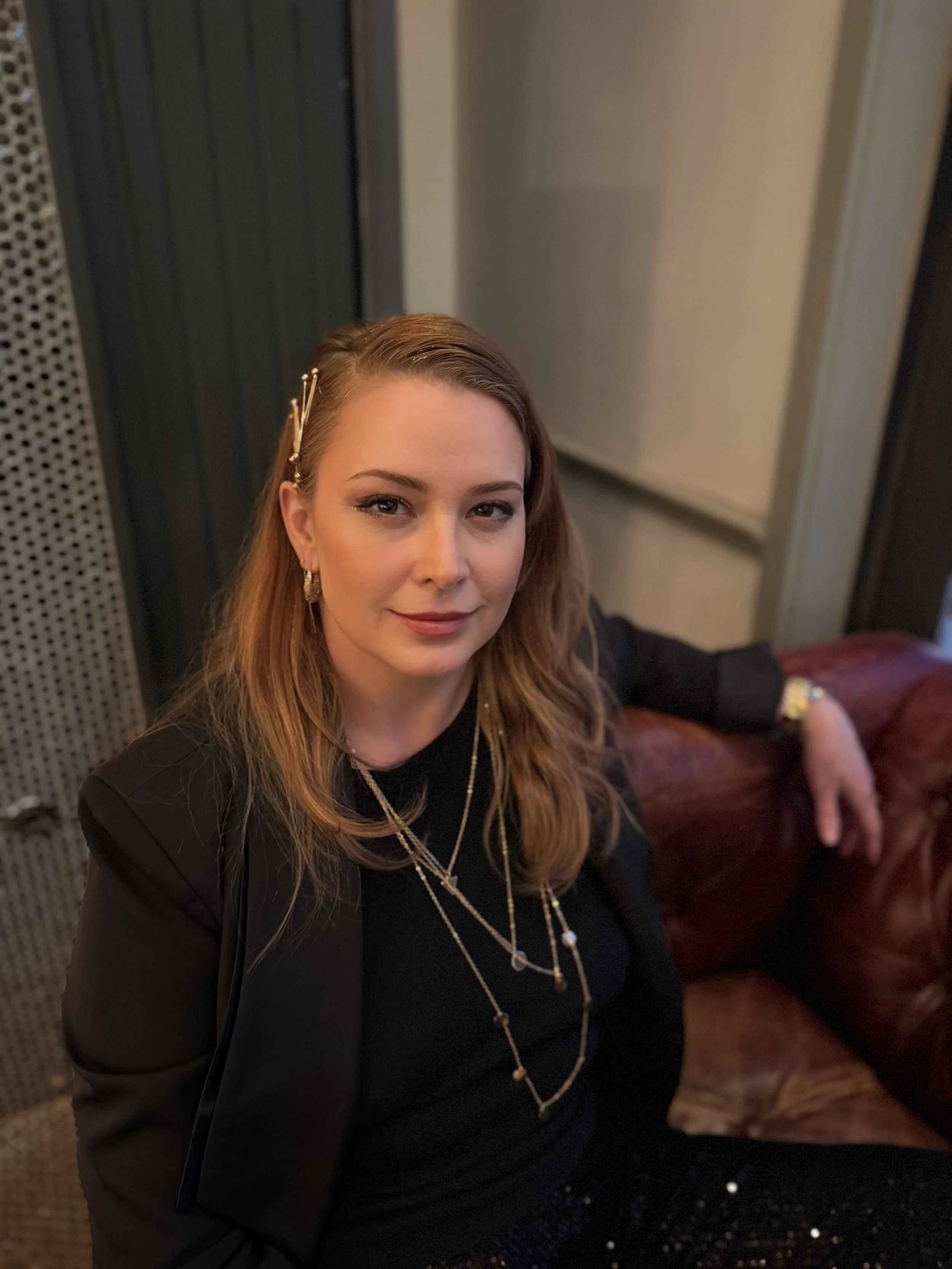 A woman with light skin, reddish-brown hair, and makeup sitting on a leather couch in a dimly lit room, wearing jewelry including earrings and necklaces, with a black blazer and blouse.