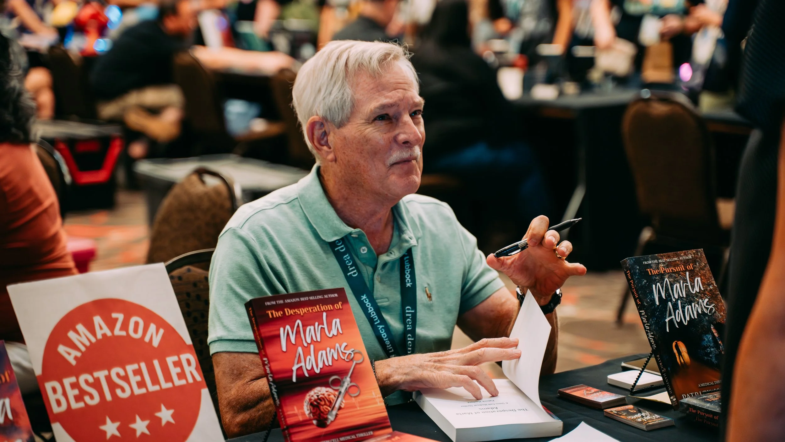 An elderly man with white hair and a mustache at a book signing event. He is wearing a light green polo shirt and has a lanyard around his neck. On the table in front of him are copies of his book, "The Pursuit of Marla Adams," a sign that says "Amazon Bestseller," and some promotional materials. The man is holding a pen and appears to be signing a book or preparing to do so, with a thoughtful expression. The background shows a crowded room with people sitting and standing, likely at a conference or literary event.