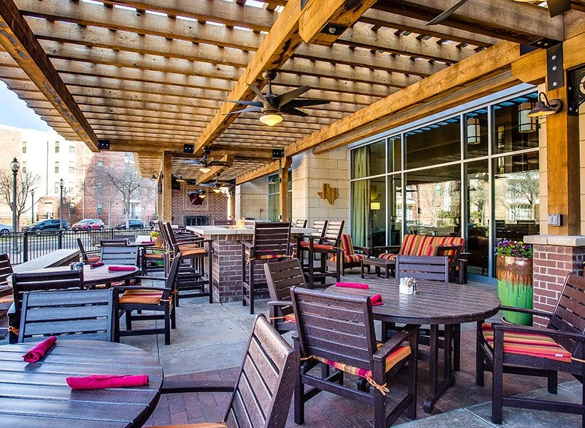 Outdoor patio of a restaurant with wooden pergola, tables, and chairs with red napkins, decorative cushions, and potted plants, surrounded by a metal fence and cityscape background.