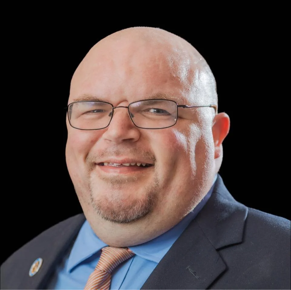 A smiling man with glasses, a bald head, wearing a suit, blue shirt, and a striped tie, against a black background.
