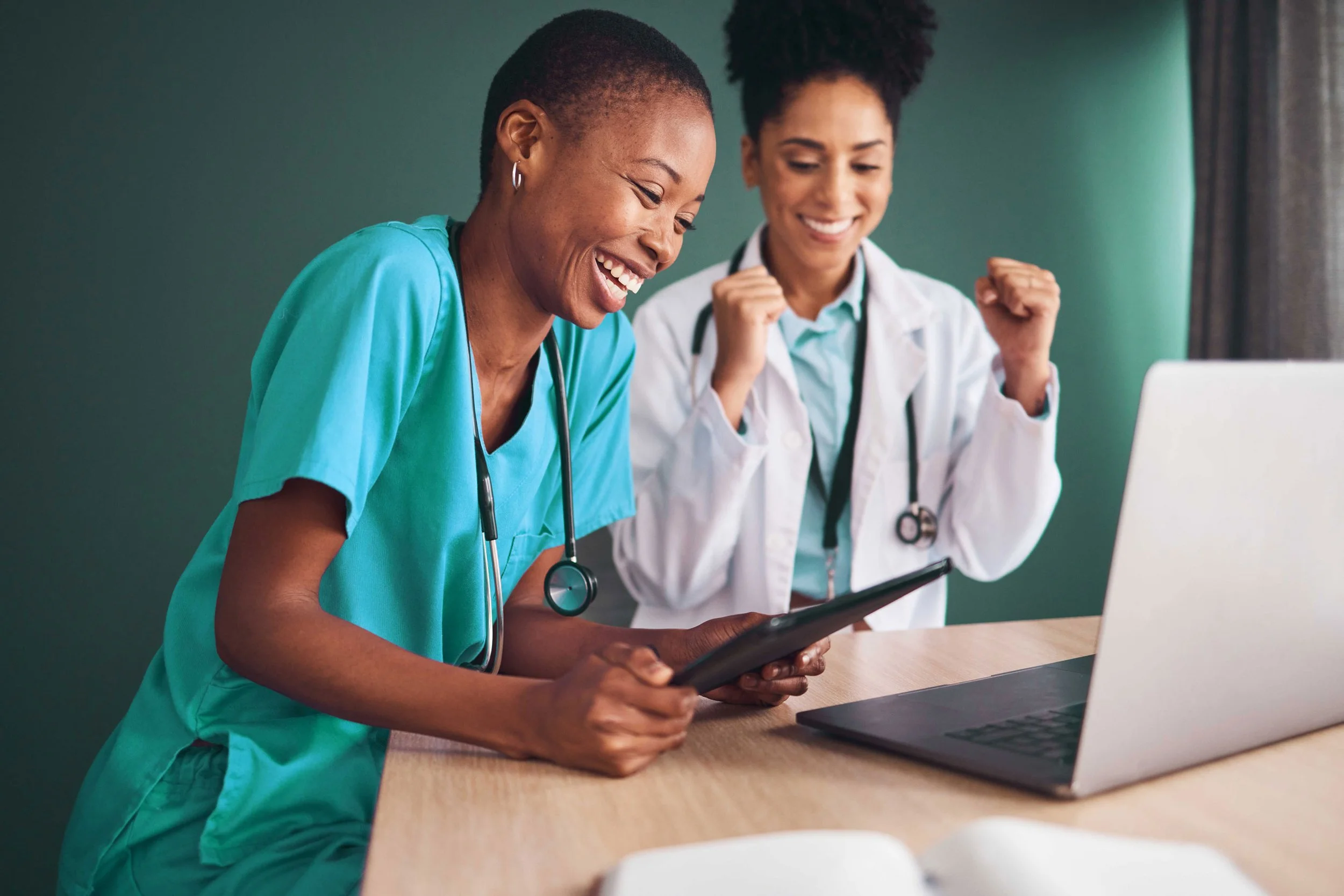 Two female healthcare professionals, one in scrubs and the other in a white coat, smiling and celebrating while looking at a tablet in a medical office.