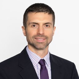 A man with short dark hair and a beard, wearing a dark suit, checkered shirt, and purple tie, smiling against a plain white background.