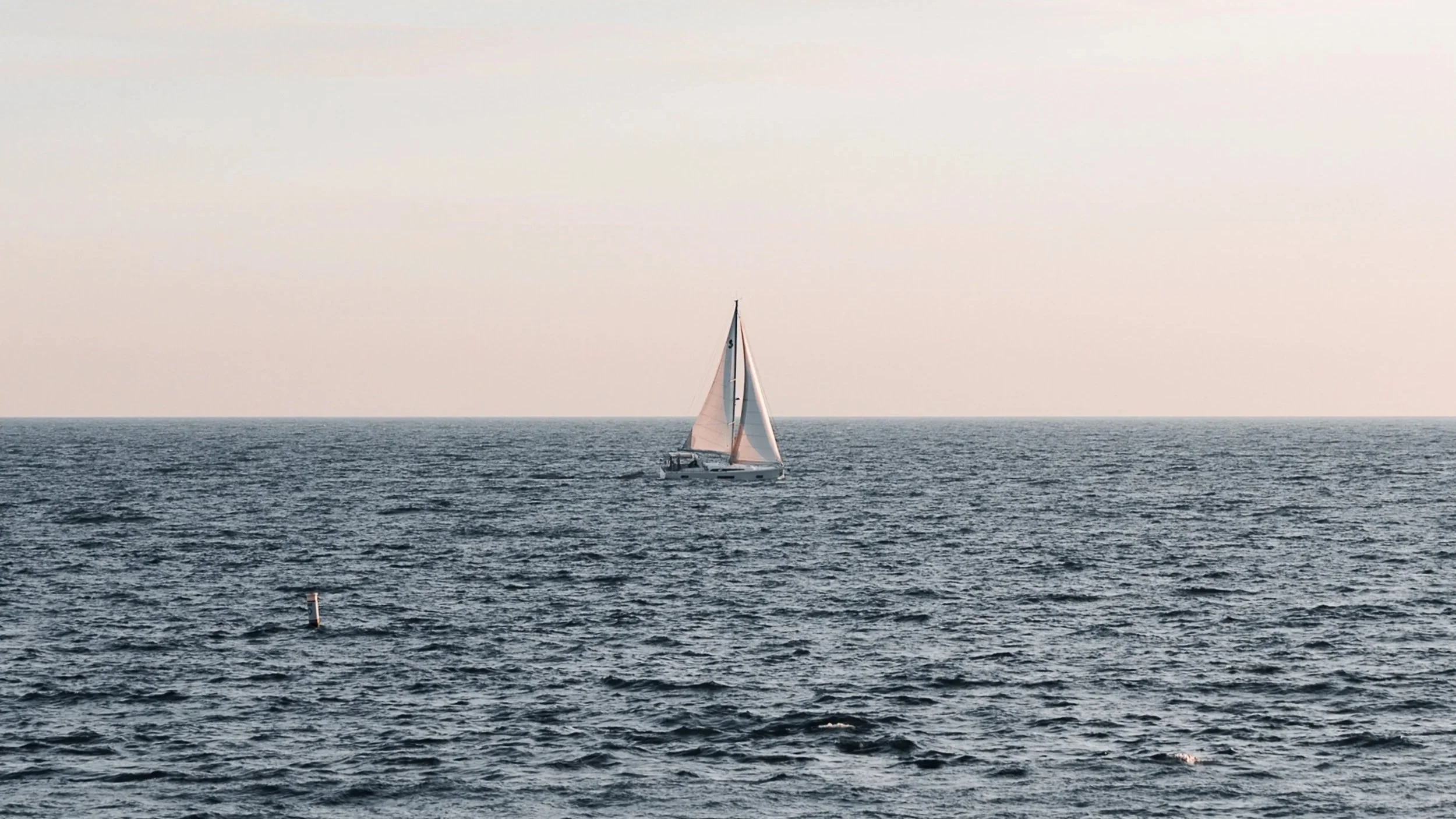 A sailboat with white sails on the open ocean during sunset.