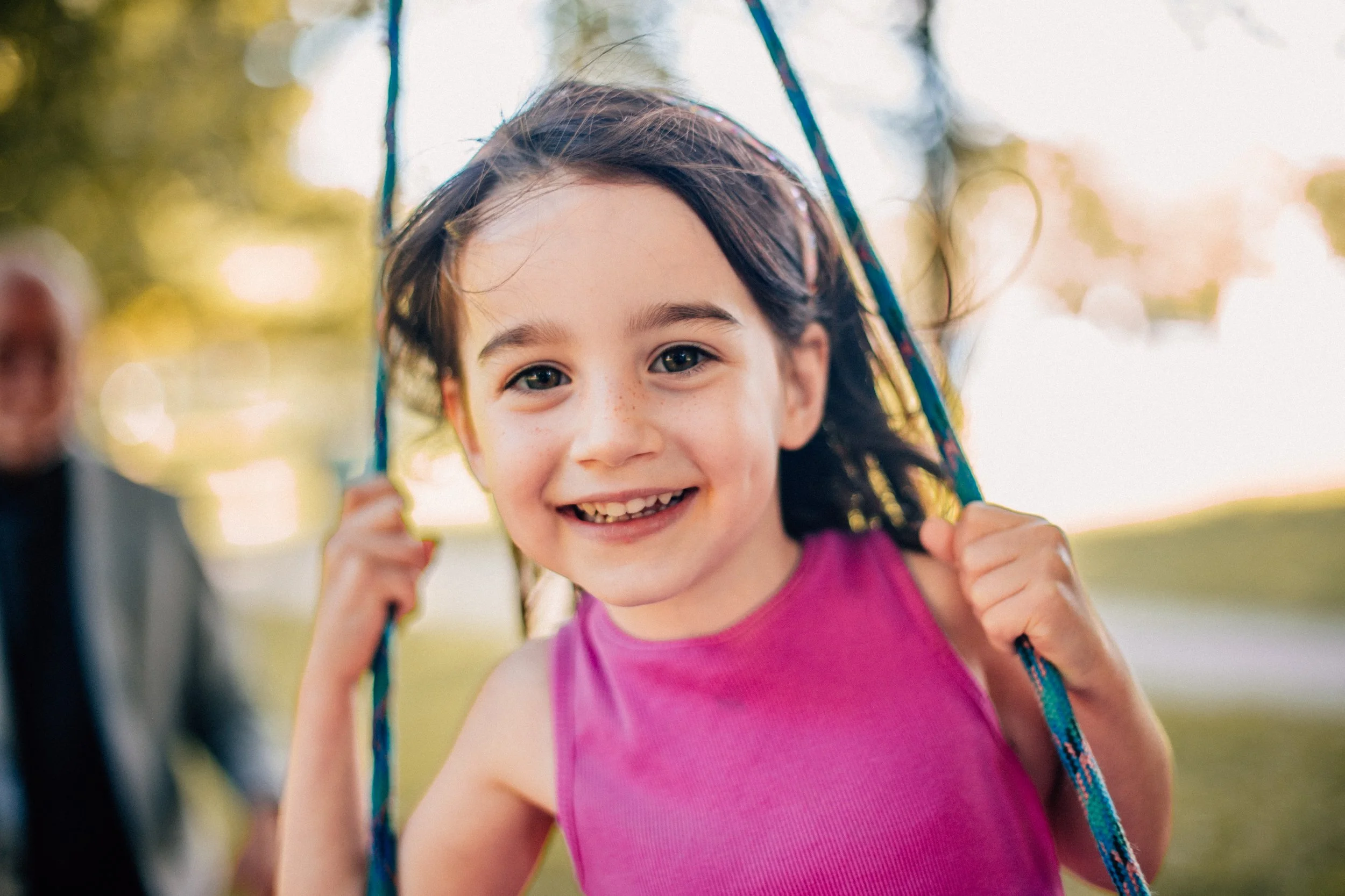 A young girl with dark hair and a pink shirt smiling while playing on a swing outdoors.