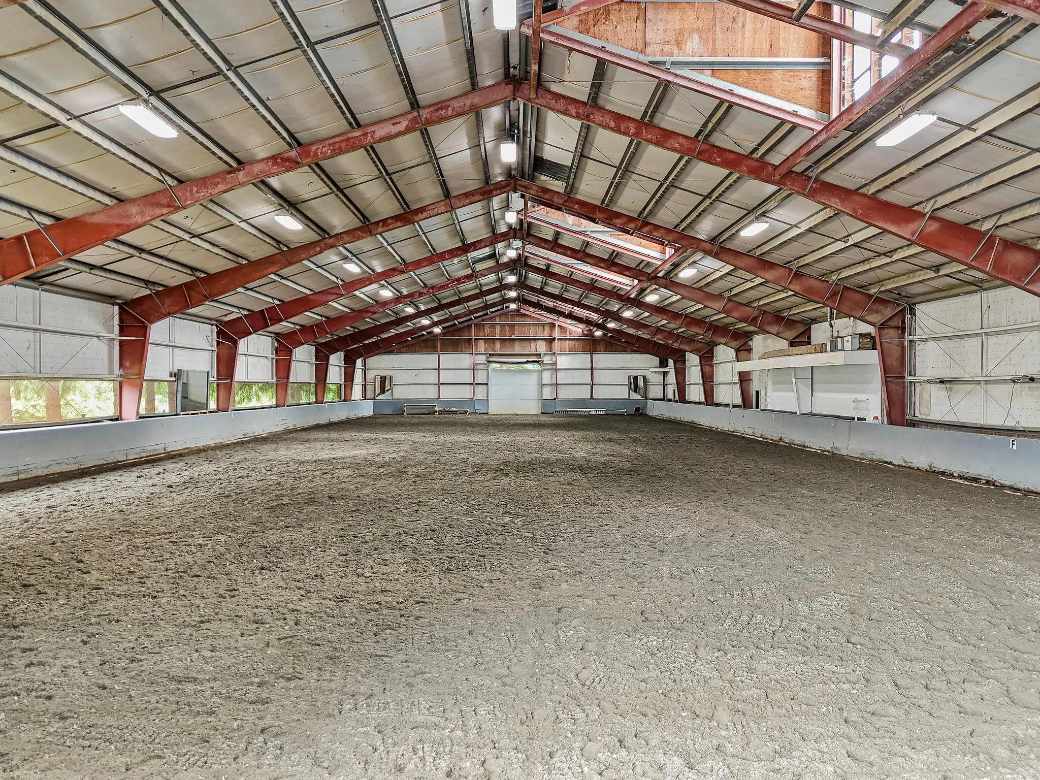 Empty indoor riding arena with dirt floor, metal structure, and windows along the side walls.