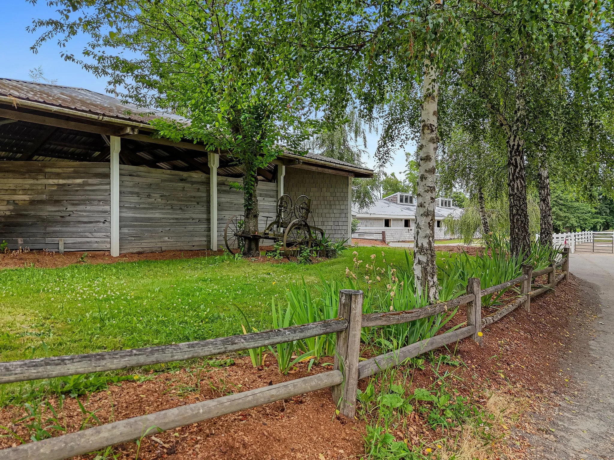 A fenced garden area with trees, green grass, and plants, next to a building with a corrugated metal roof, and a dirt pathway to the right.