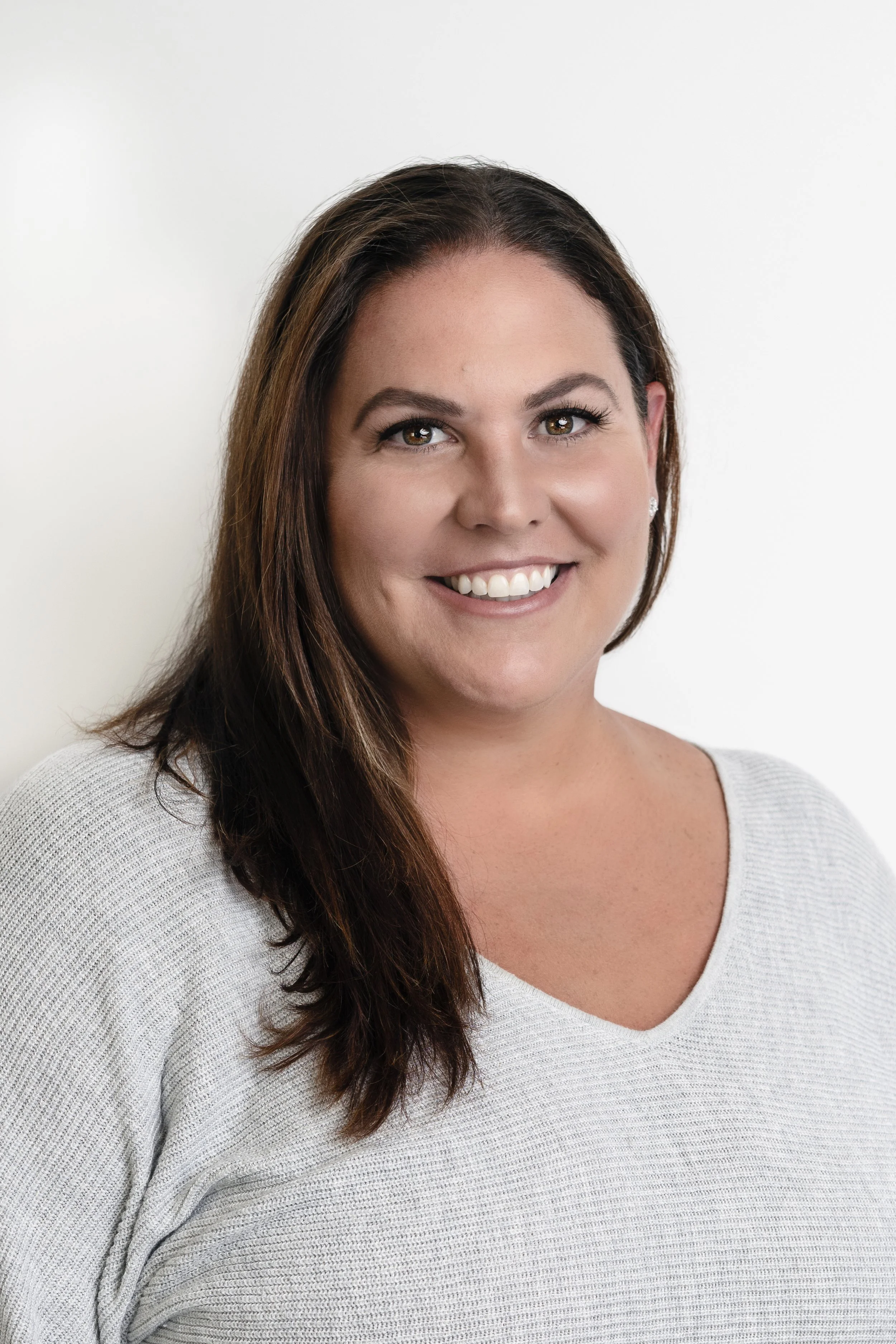 A woman with long brown hair and light makeup, wearing a light gray sweater, smiling in front of a white background.