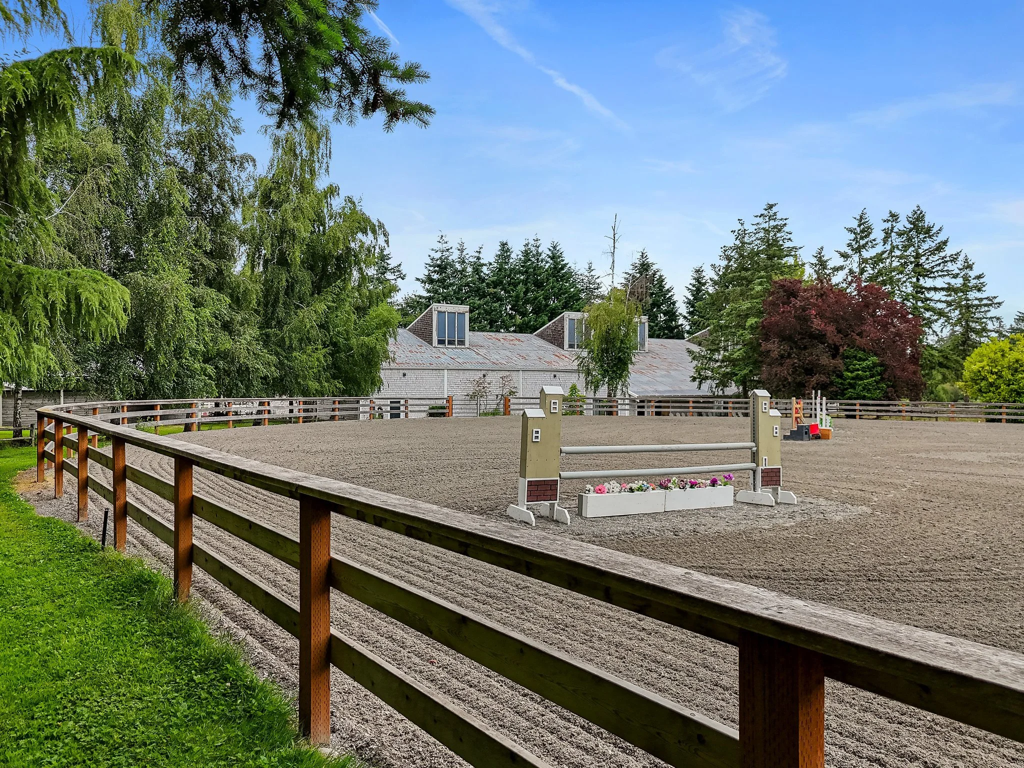 An outdoor riding arena with a dirt surface, surrounded by a wooden fence, with trees and buildings in the background under a partly cloudy sky.