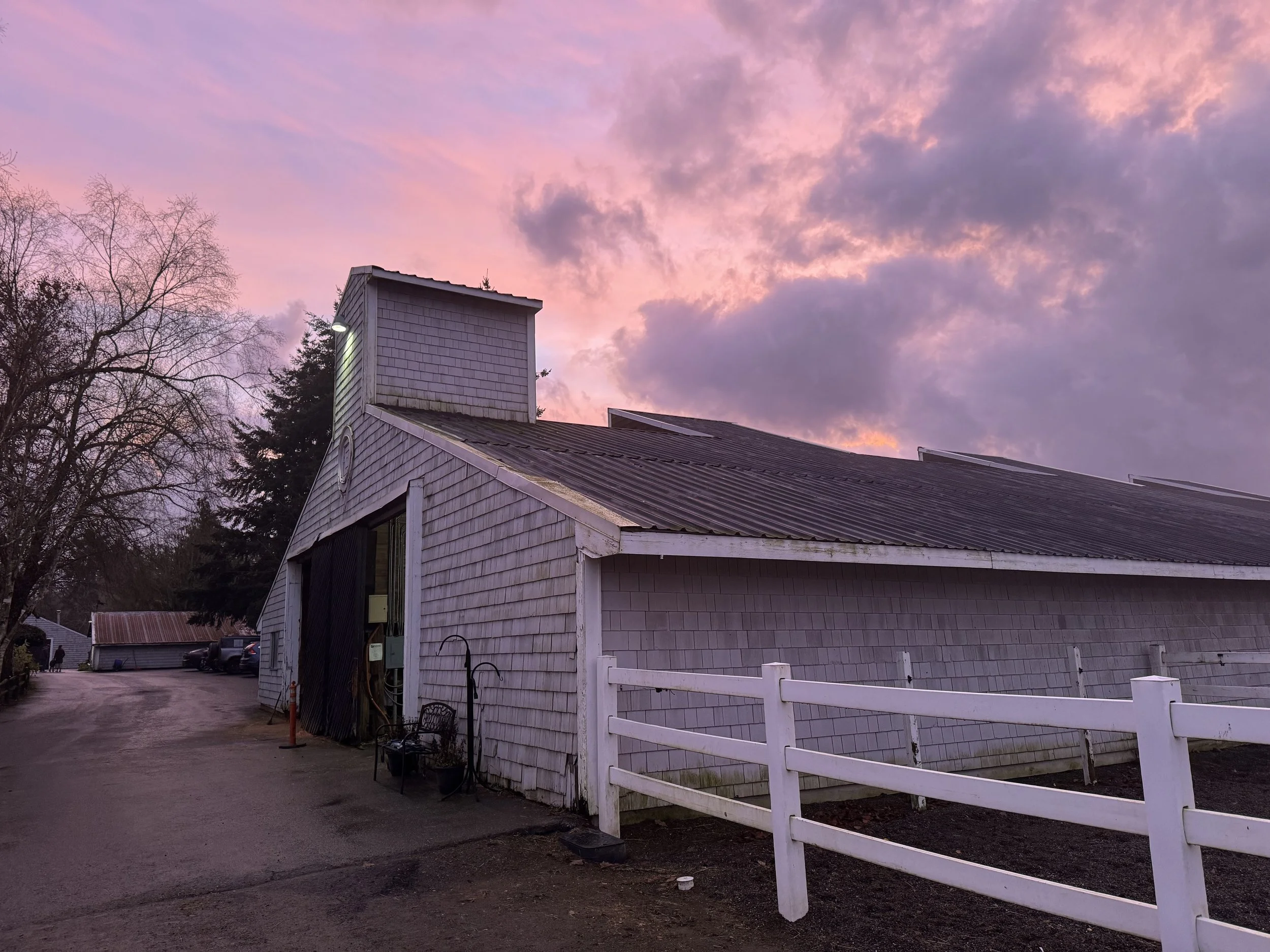 A white building with a sloped roof and shingle siding, featuring a small tower-like structure on top, under a colorful sky at dusk with pink and purple clouds. There is a white fence along the side of the building and several trees in the background