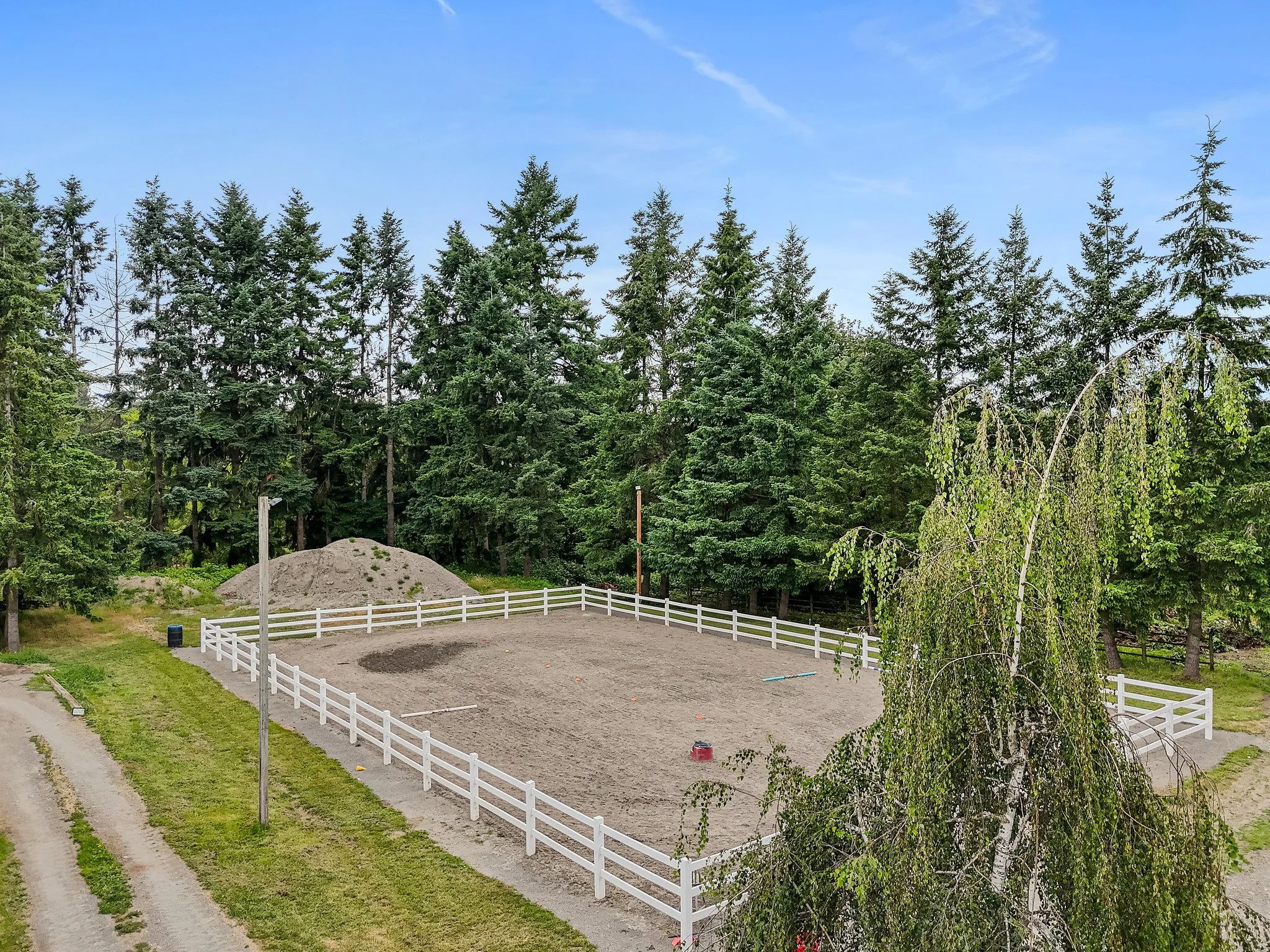 An outdoor riding arena enclosed with a white fence, with a dirt and sand mixture ground surface, trees surrounding it, and a mound of dirt in the background. There are a few small objects and equipment inside the arena.