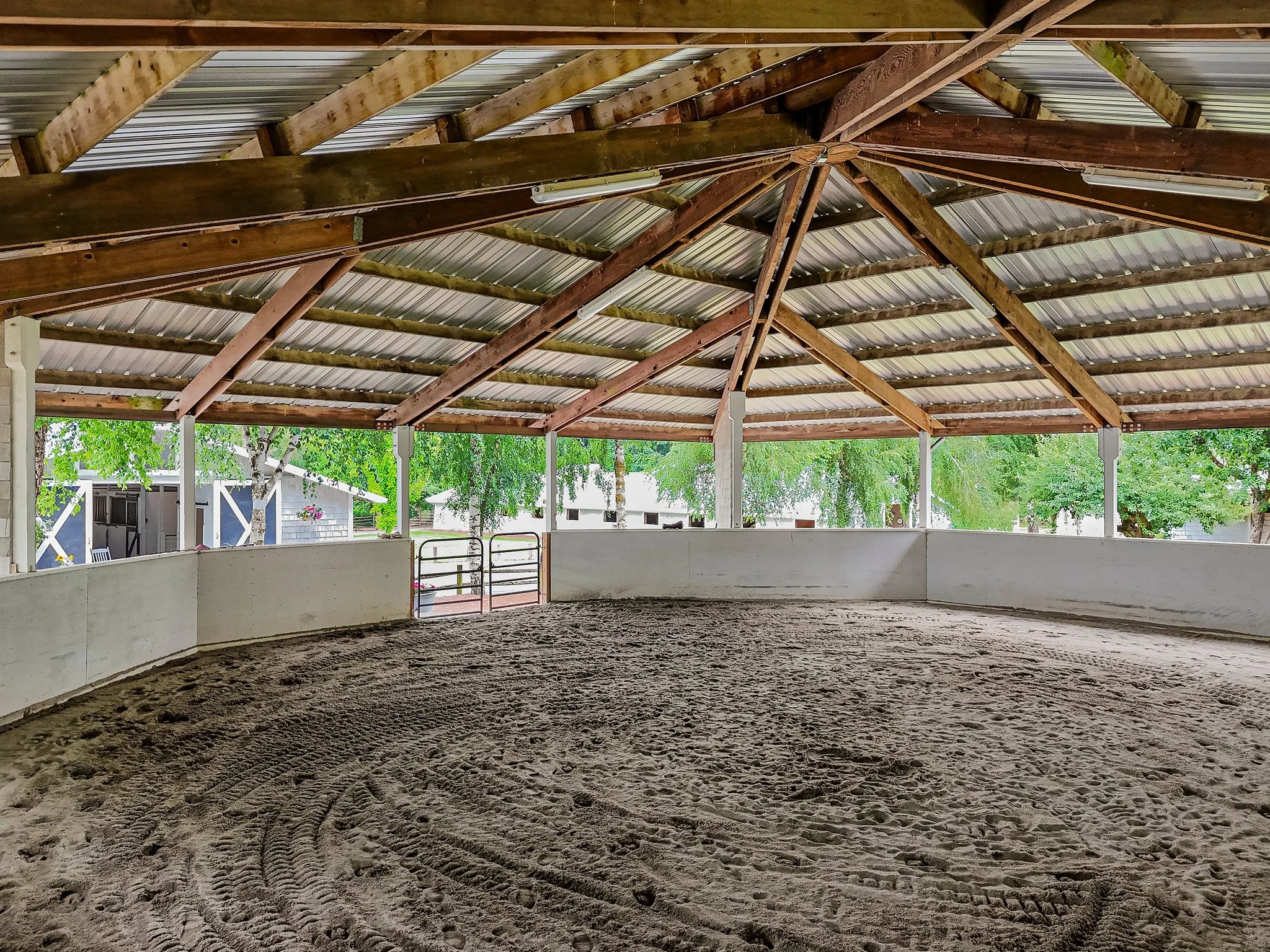 Indoor riding arena with sandy footing, surrounded by white walls, wooden roof structure, and open sides with trees visible outside.