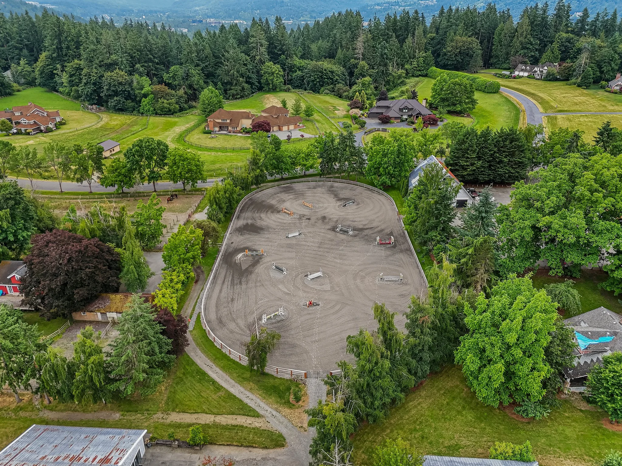 An aerial view of an outdoor horse riding arena surrounded by green trees and residential houses in a rural area.