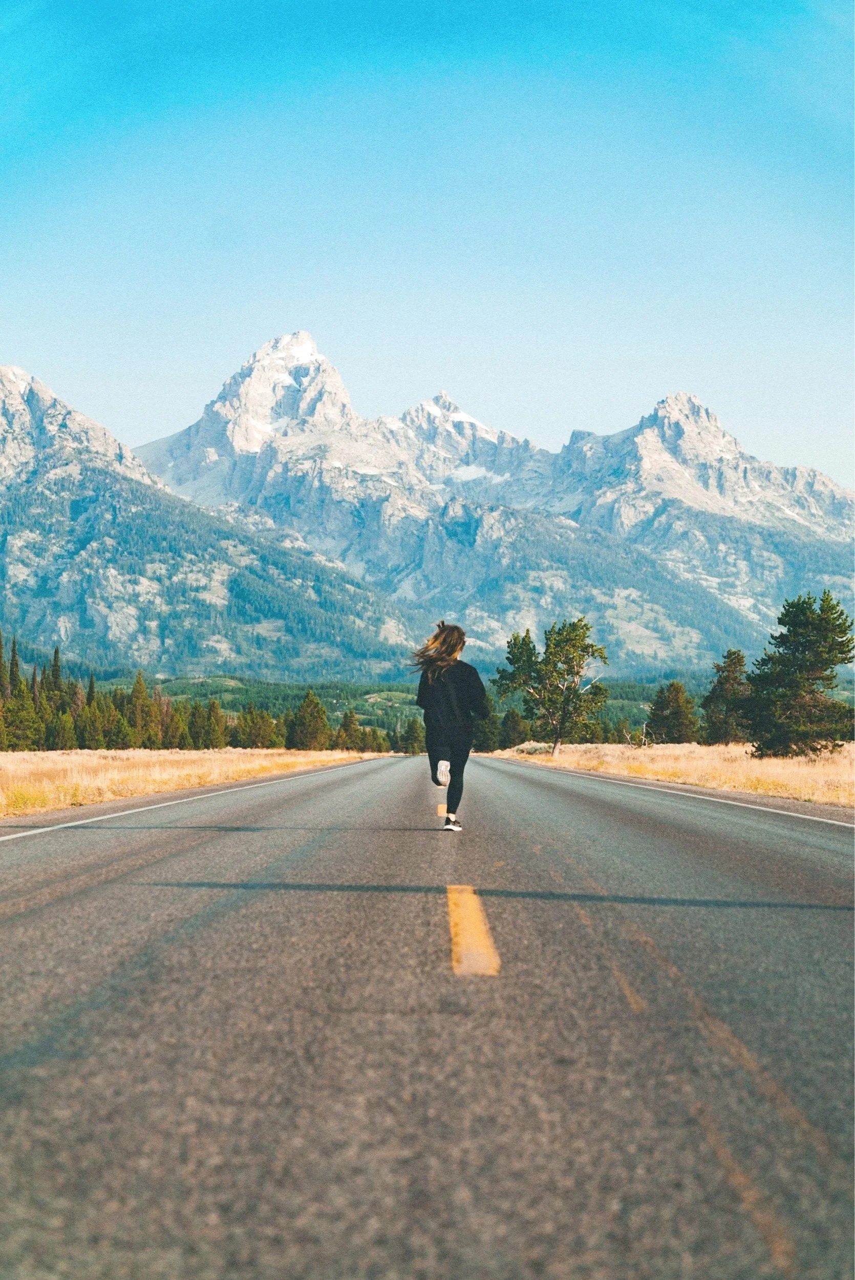 A person walking down a deserted road with mountains in the background and a clear blue sky.