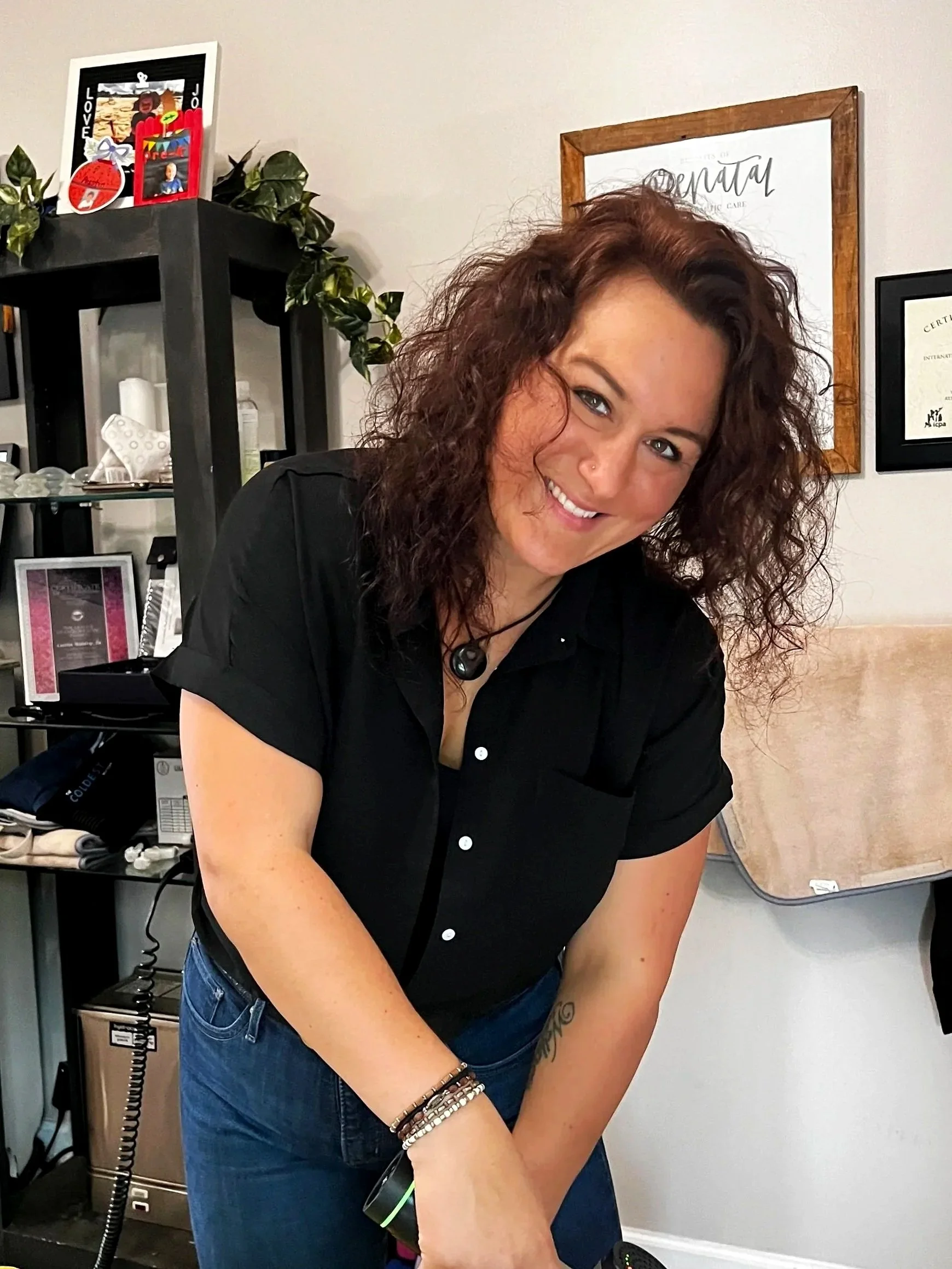 A woman with curly brown hair, smiling, wearing a black blouse and jeans, leaning forward in an indoor setting with framed certificates and decorations in the background.