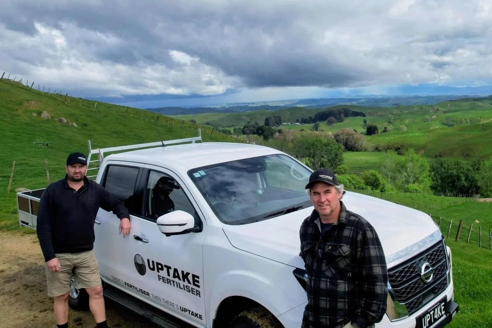 Two men standing next to a white pickup truck with 'UPTAKE FERTILISER' written on the side, in a green rural landscape with rolling hills and cloudy sky.