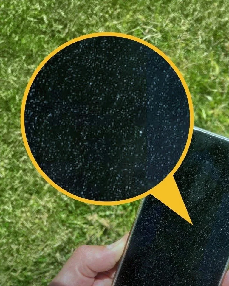 Close-up of a smartphone screen with many small white spots, held outdoors above green grass.