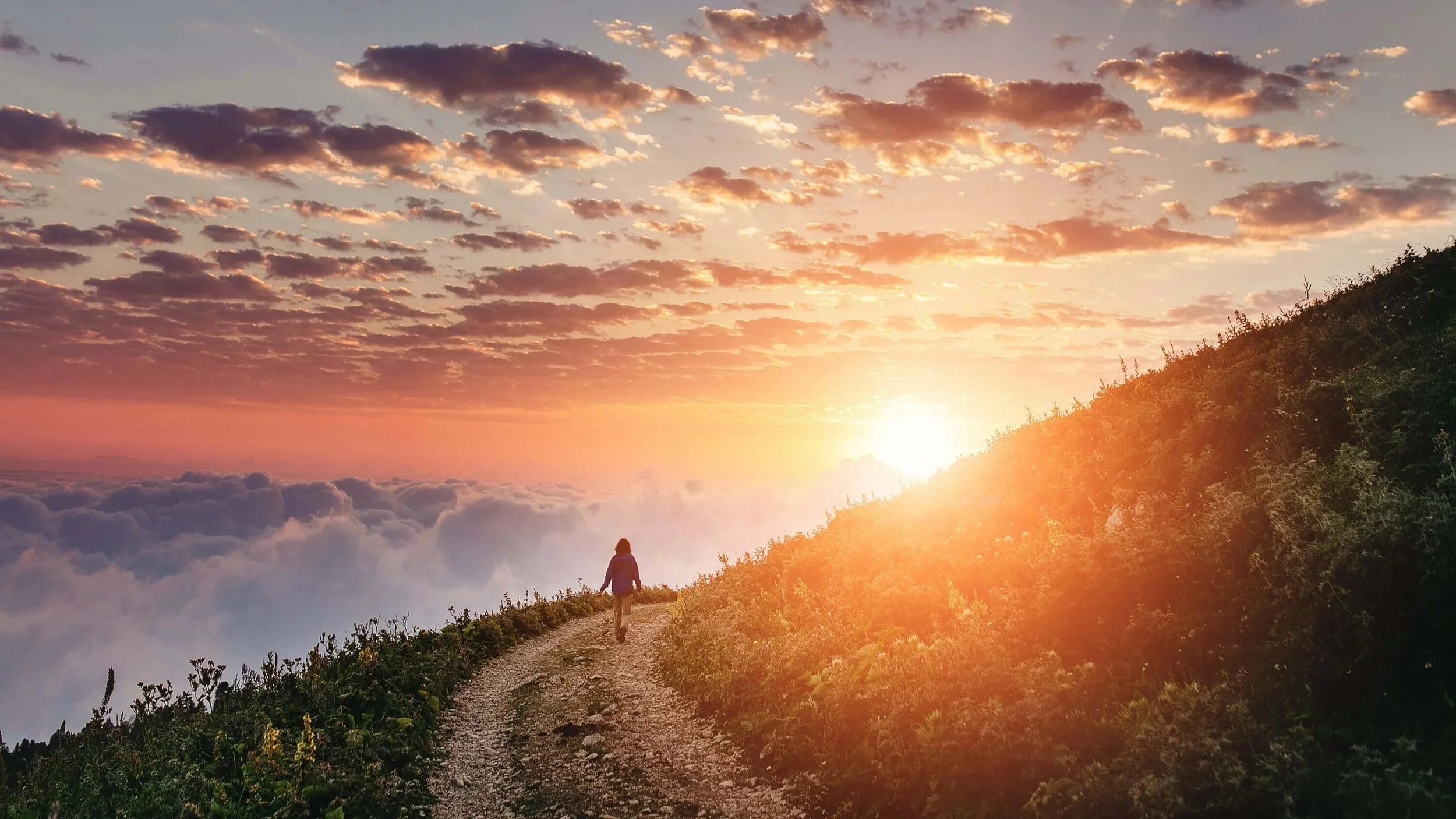 A person walking along a rocky dirt road, high above the clouds, towards a sunrise