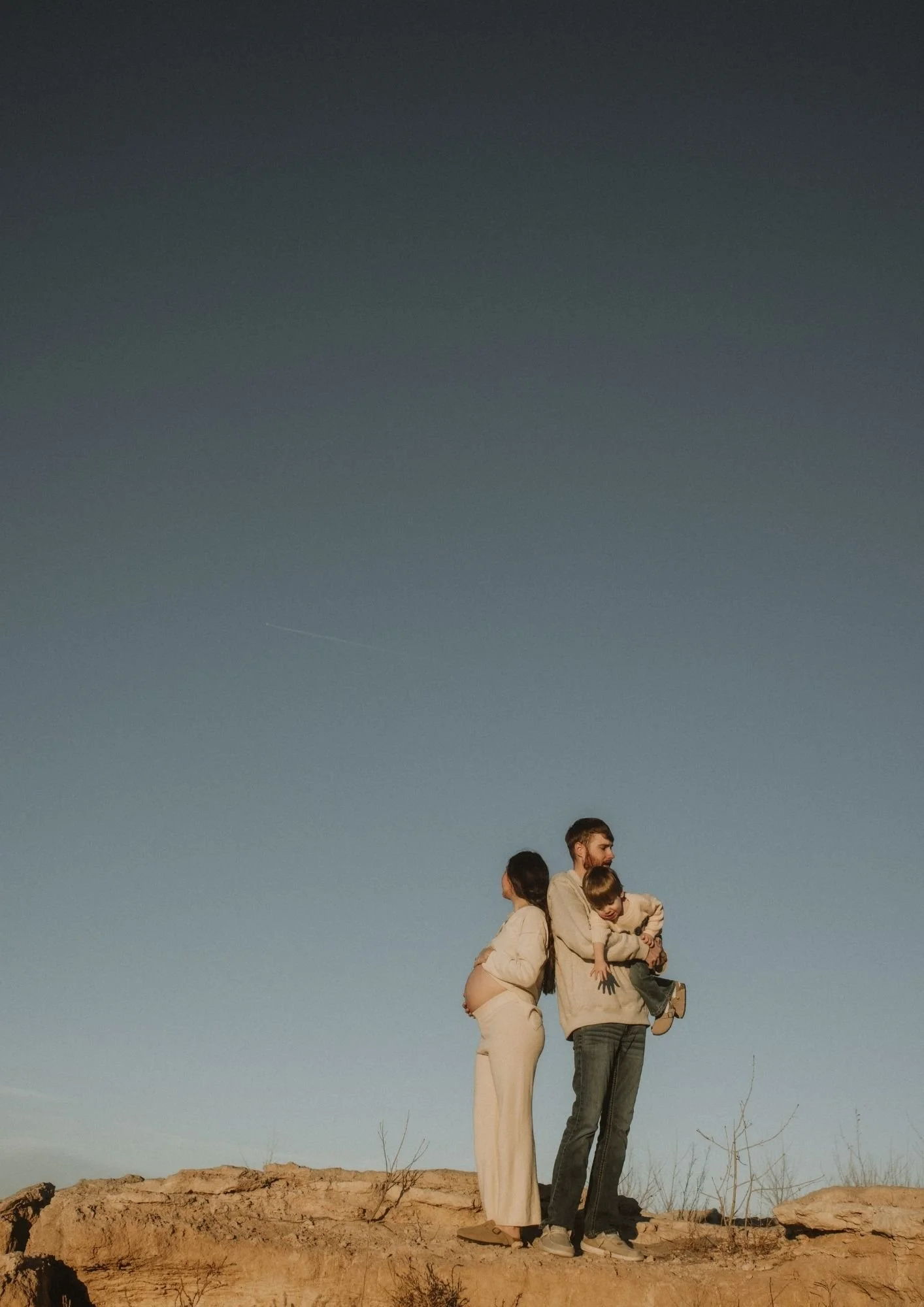 A family standing on rocky terrain under a clear evening sky. The pregnant woman stands to the left, the man holds a child in his arms to the right, and they are all facing different directions.