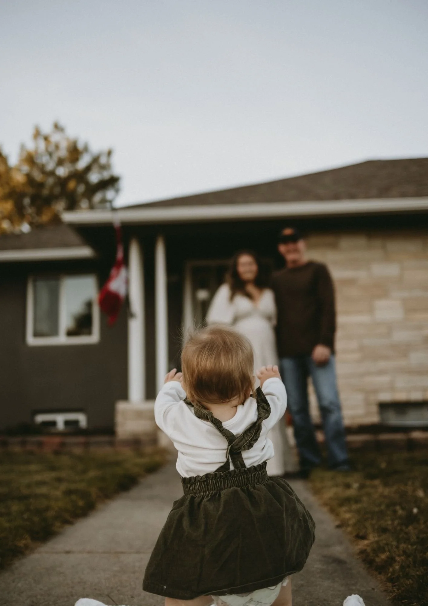 A young child crawling on a sidewalk towards a house, with a couple standing in front of the house blurred in the background.