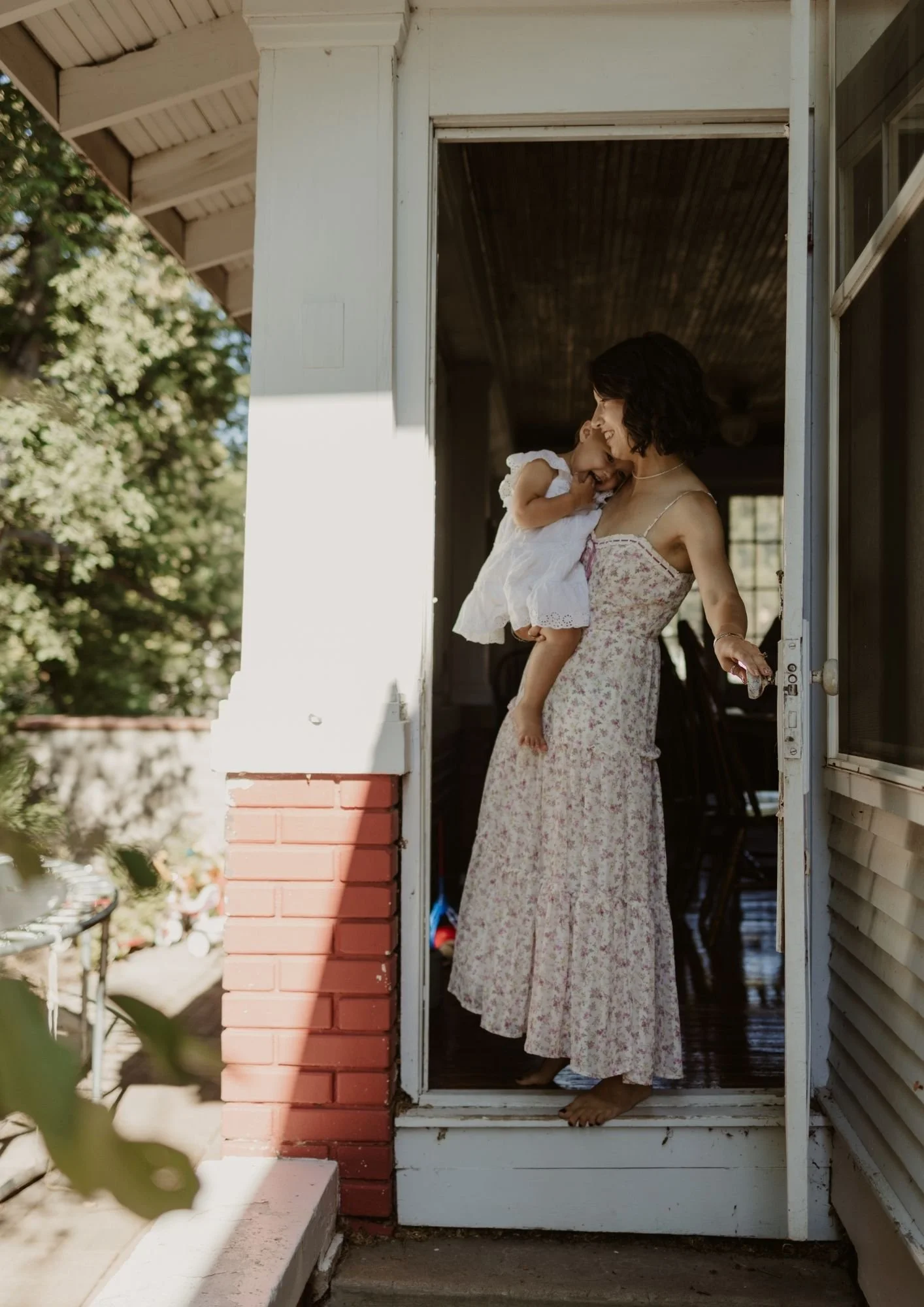 A woman holding a young girl on a porch, smiling and interacting with her inside a house during daytime.