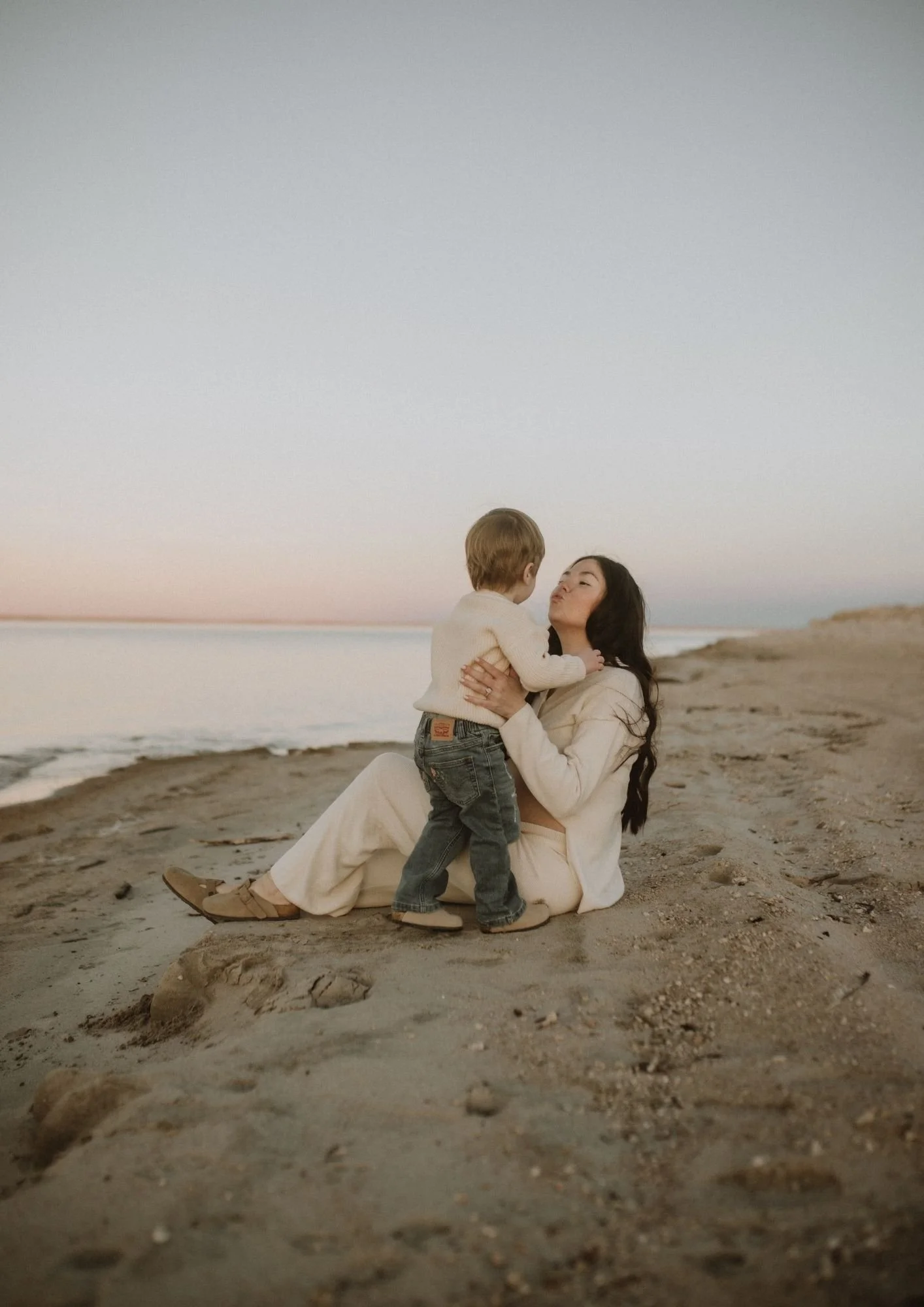 A woman sitting on a sandy beach holding a young child, sharing a kiss as the sun sets, with a calm ocean in the background.