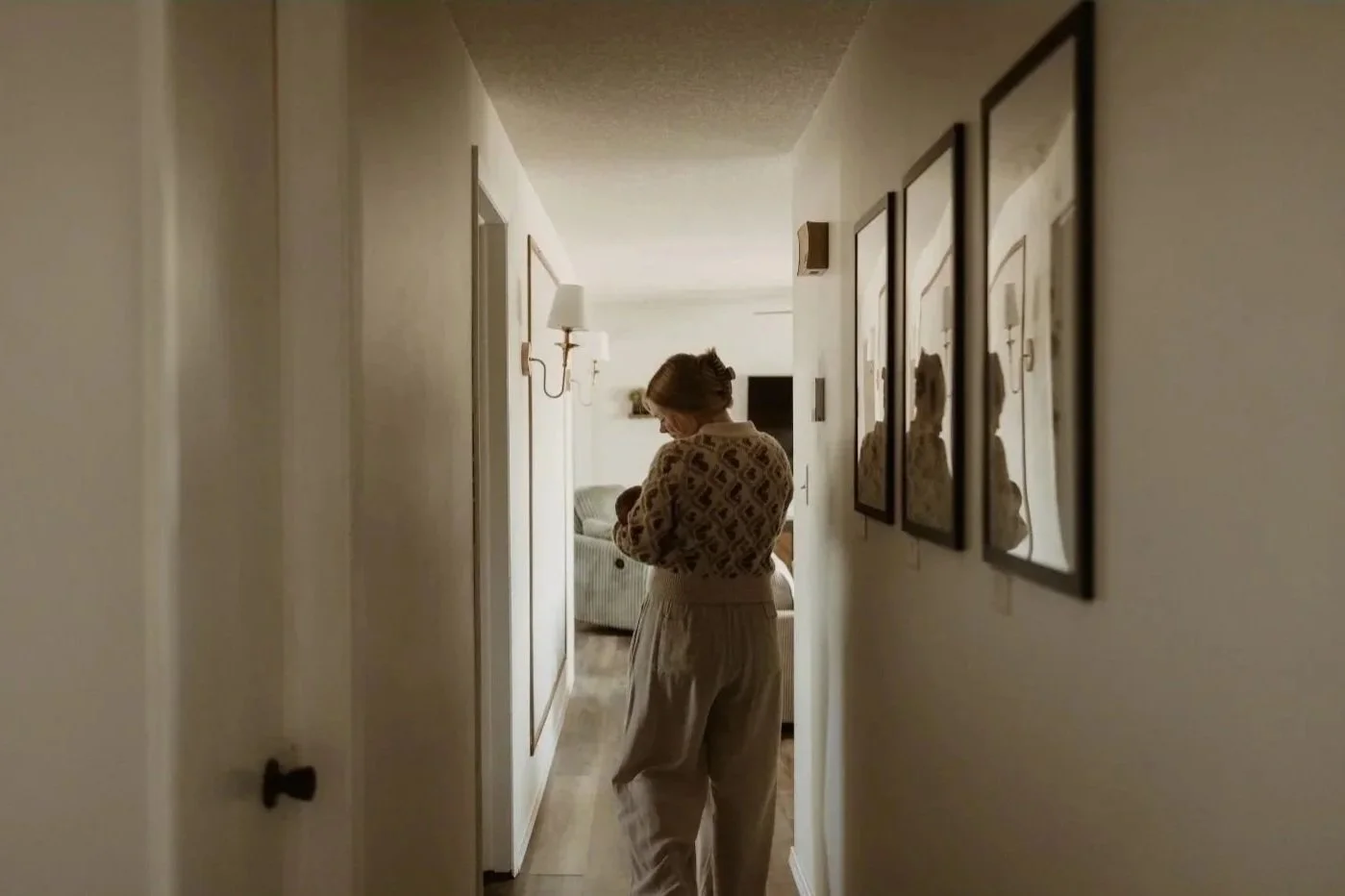 A woman holding a baby in a hallway with framed pictures on the wall and a light fixture, leading to a living room with a couch.