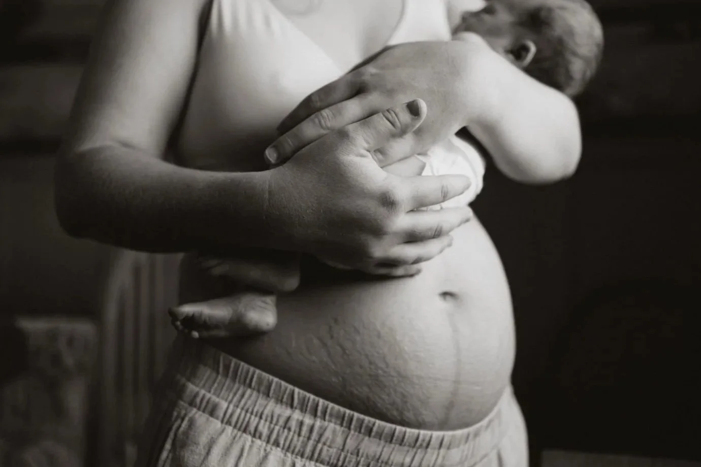 A person with a bare midriff wearing light-colored shorts is holding their chest with both hands, embracing a young girl who is leaning against them. The image is black and white, showing the person's torso and the girl resting her head on the person