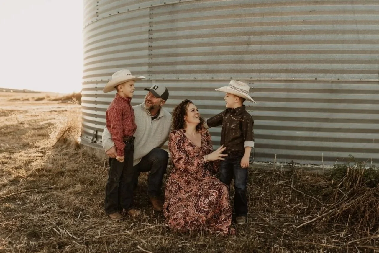 A family of four, including two children with cowboy hats, standing outdoors near a large metal grain silo during sunset, smiling and interacting with each other.