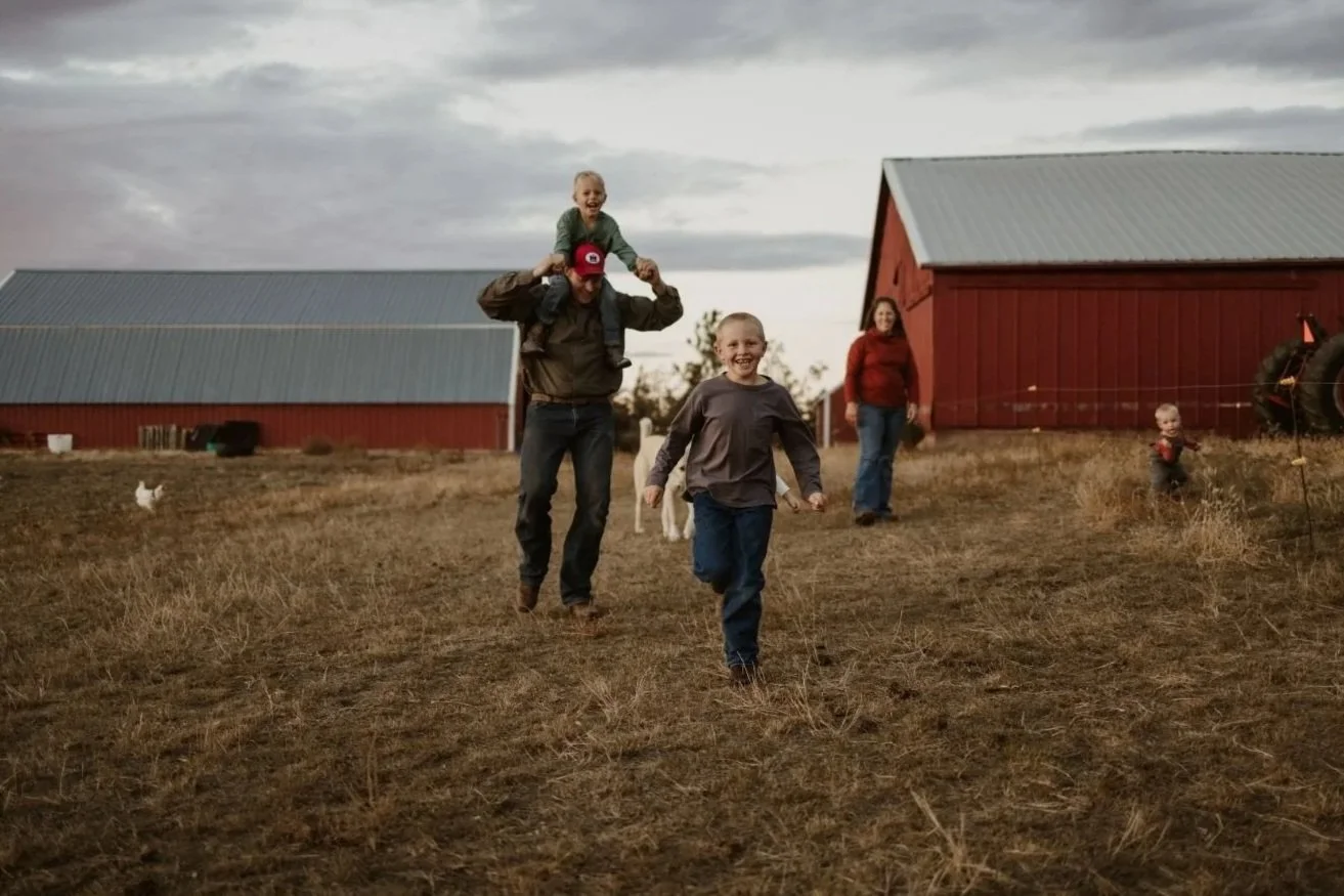 Family on a farm with red barns, children playing and adults walking on a dirt field during cloudy weather.