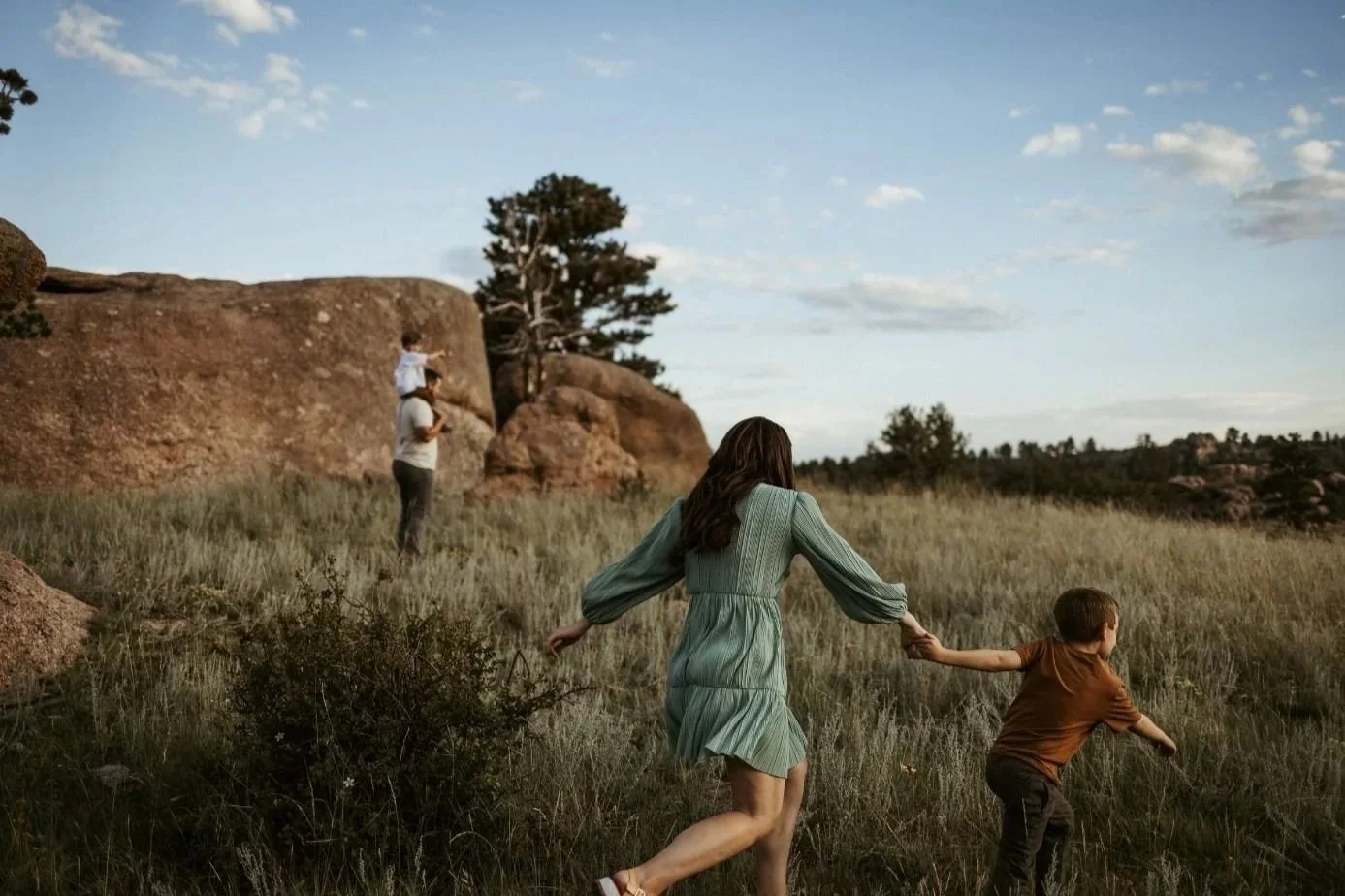 A woman and a young boy holding hands running through a grassy field with rocks and trees in the background under a cloudy sky.