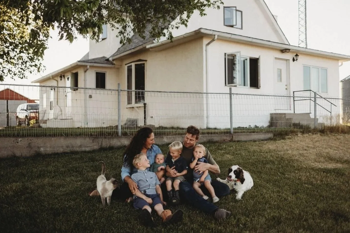 A family with three children and two dogs sitting on a lawn in front of a house, smiling and spending time together in the evening.