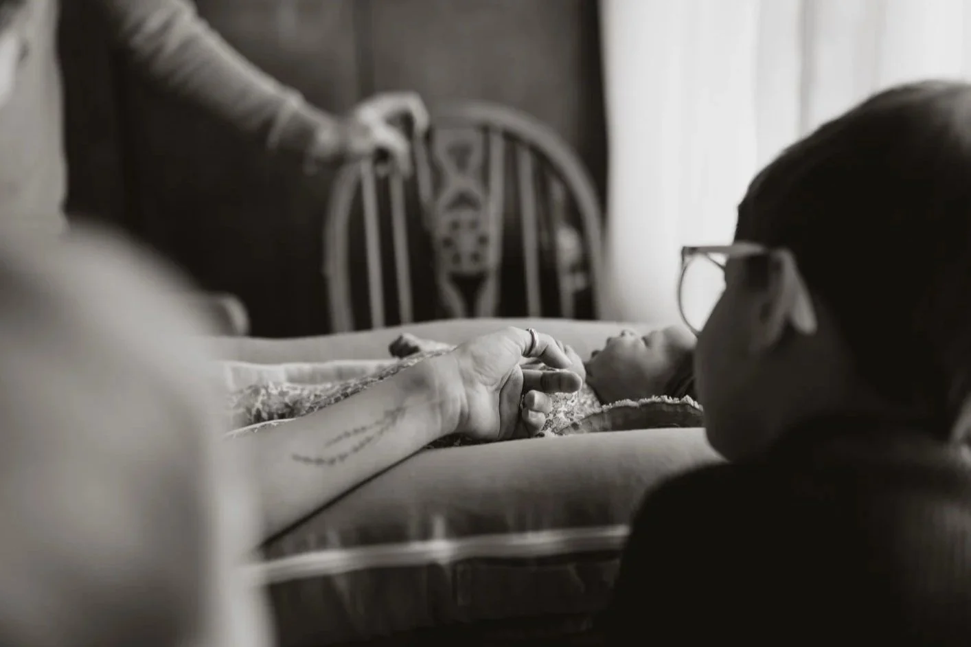 A person lying in a hospital bed holding hands with another person who is sitting beside them, with a wheelchair visible in the background.