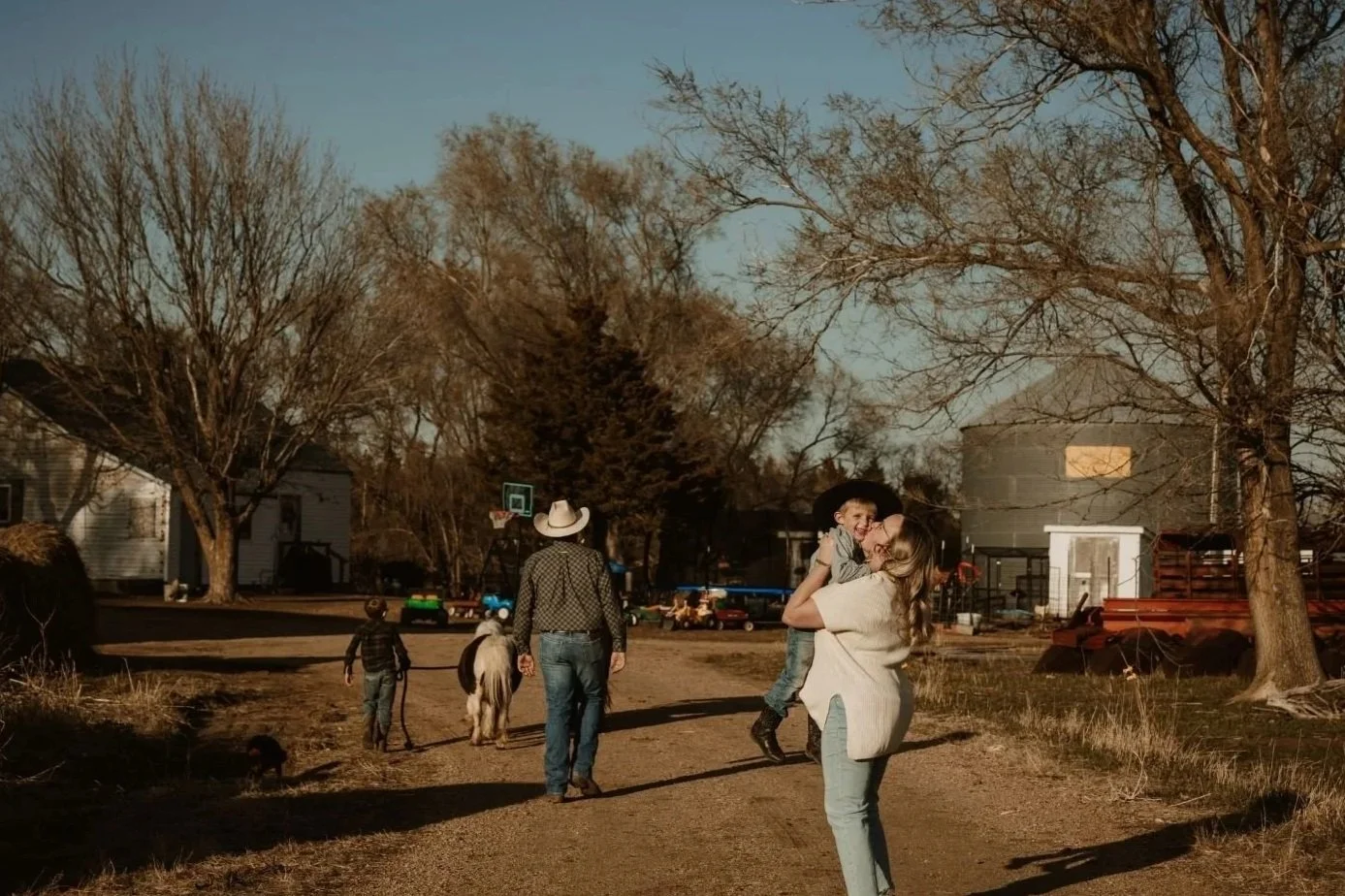A woman holding a young child in a joyful embrace outdoors during daytime, with a woman wearing a cowboy hat, a child, and a pony on a dirt path, surrounded by leafless trees and residential buildings.
